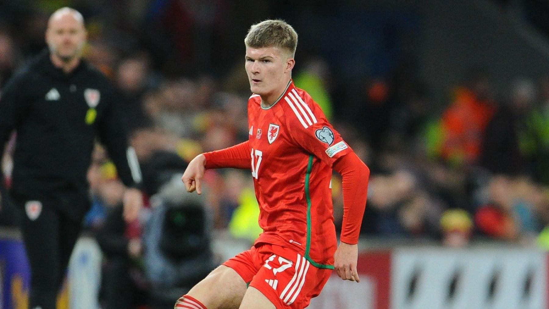 Jordan James, playing for Wales, wears their red kit and looks at the ball, off camera, with his right arm raised halfway up his side and his left hanging at his hip. 