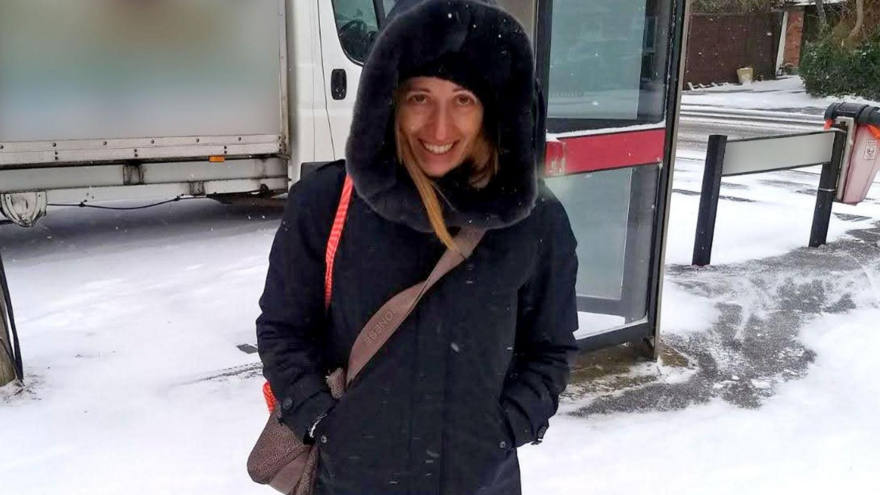 A woman in a navy blue warm coat, with the hood up, is standing in snow with a brown across-body bag. Behind her is a van, a phone box and a street sign that's blurred out.