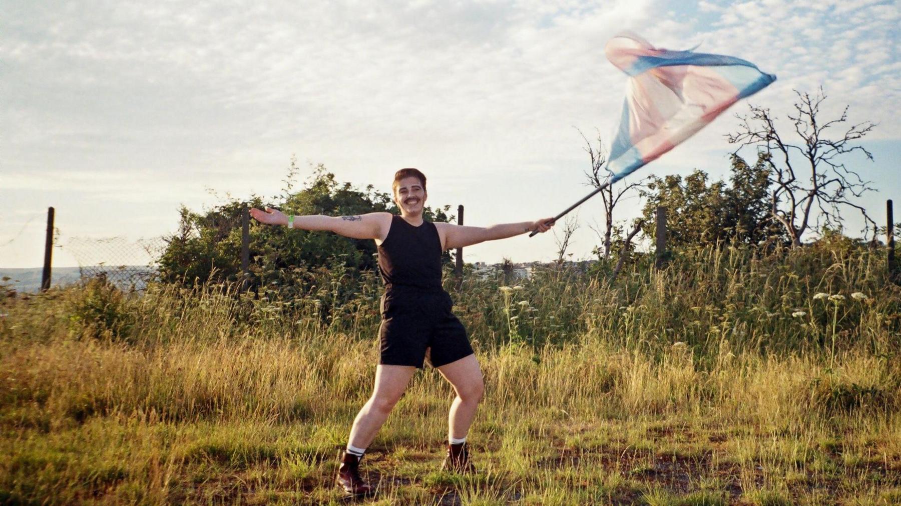 A person in a black vest and shorts stands in a field waving a trans flag.