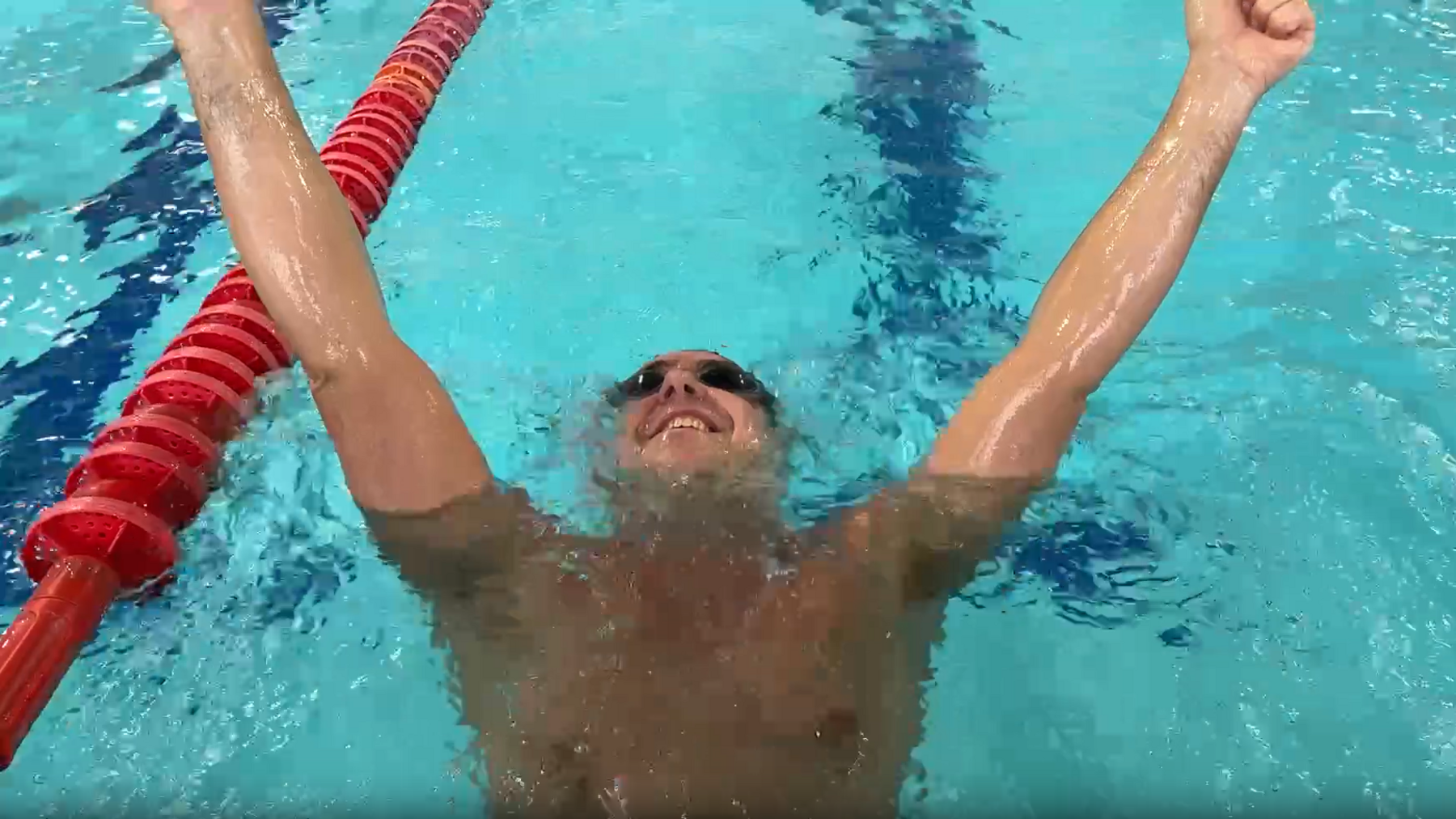 Edward Sault in the lane of a swimming pool with his arms in the air in celebration. He is wearing black goggles.