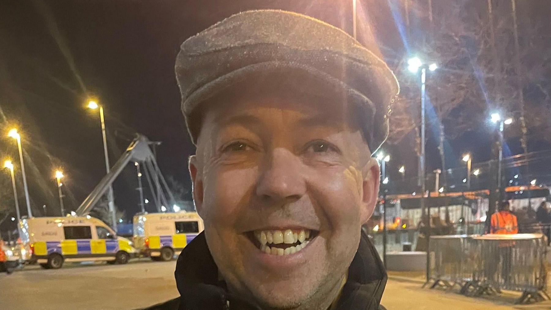 Plymouth Argyle fan Steve smiles as he leaves the Etihad Stadium in Manchester. He is wearing a grey flat cap. Two police vans are behind him and a safety steward is walking past some fencing over Steve's right shoulder. It is night time and several street lights are on.