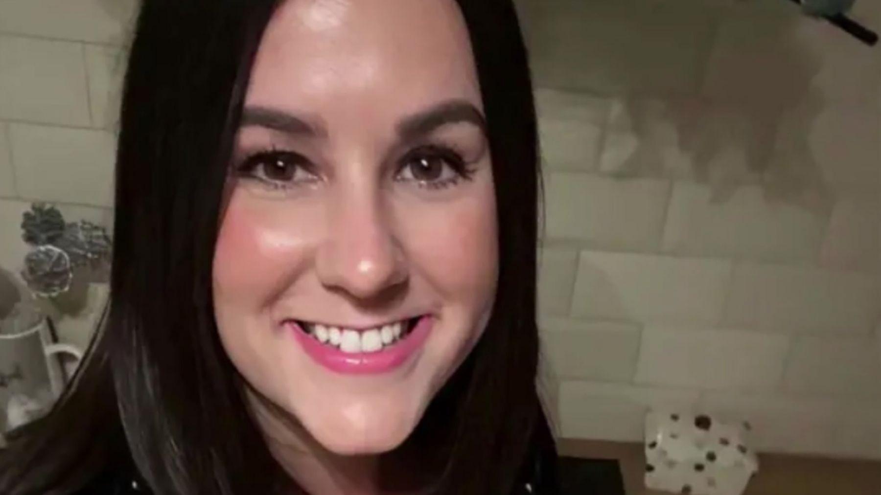 A smiling Lucy Connolly looking directly down the camera. She has shoulder length, dark brown hair and brown eyes. She is seen indoors, in what appears to be a kitchen, with a white tiled splashback and work surface behind her. The edge of a hob extractor fan can also be seen behind her.