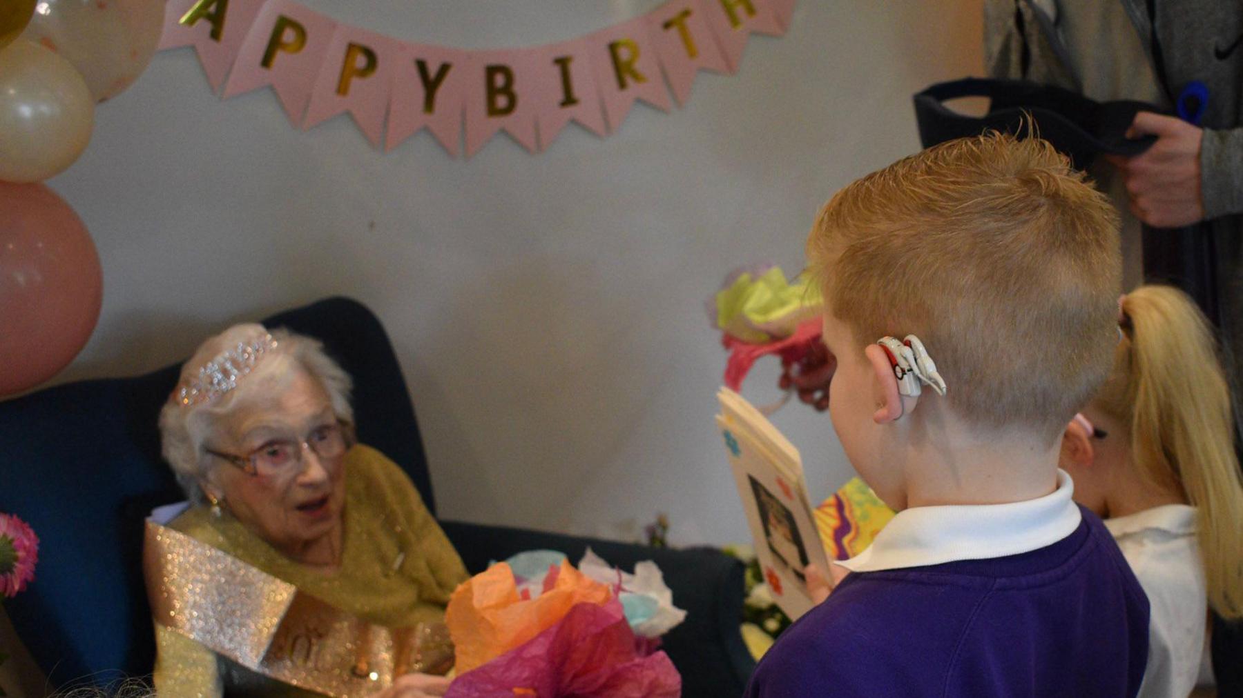 Maisie Dalziel, who is wearing a sparkly sash and a tiara, looks pleasantly surprised as two children hand her gifts and cards.