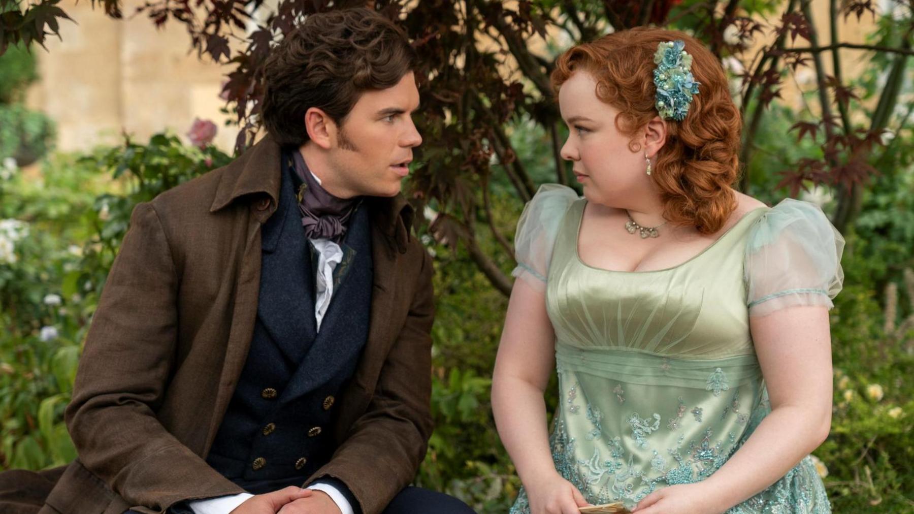 Luke Newton as Colin Bridgerton and Nicola Coughlan as Penelope Featherington sitting on a bench