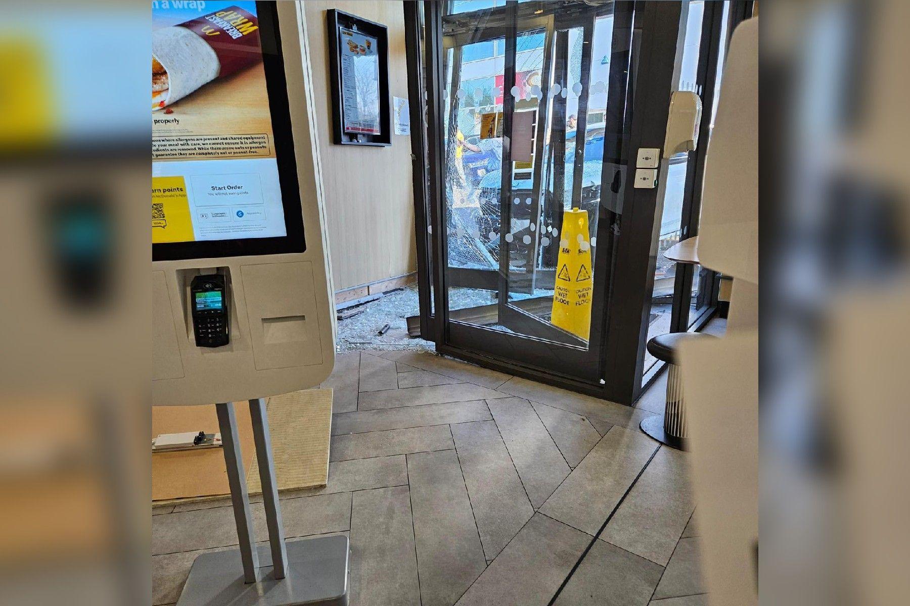 A self-service machine is in the left of the image, glass doors with dark frames are towards the middle of the image. Broken glass lays on the floor. A white car is immediately the other side of the glass doors.