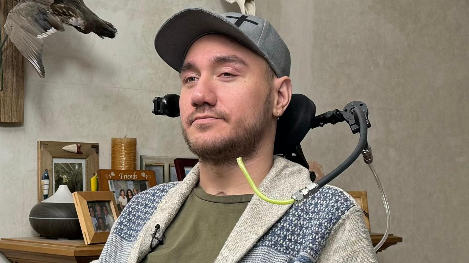 Noland. He is sitting in a wheelchair and has a tube that can be blown into to operate it. He is sharply dressed with a green shirt, white jacket and a grey baseball cap. He has well kept short hair and stubble.