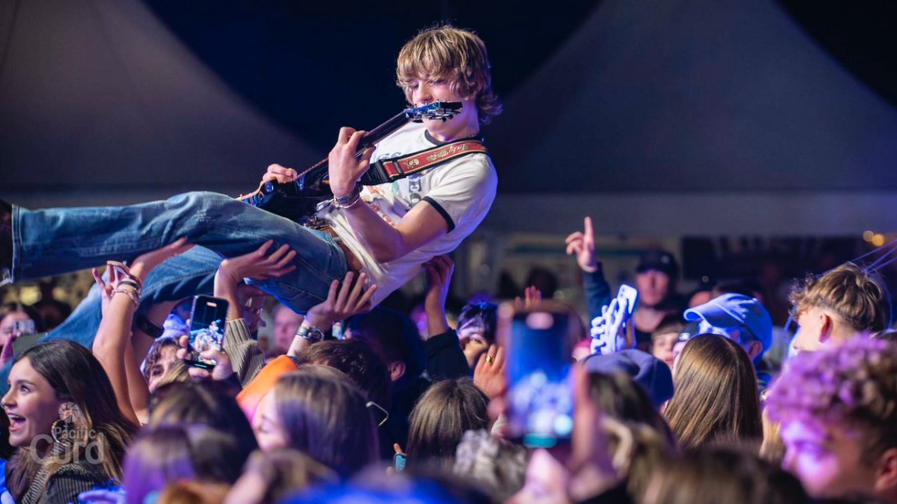 A man wearing jeans and a white t-shirt is crowd surfing while playing the guitar. Some people in the crowd are filming him on their phones.