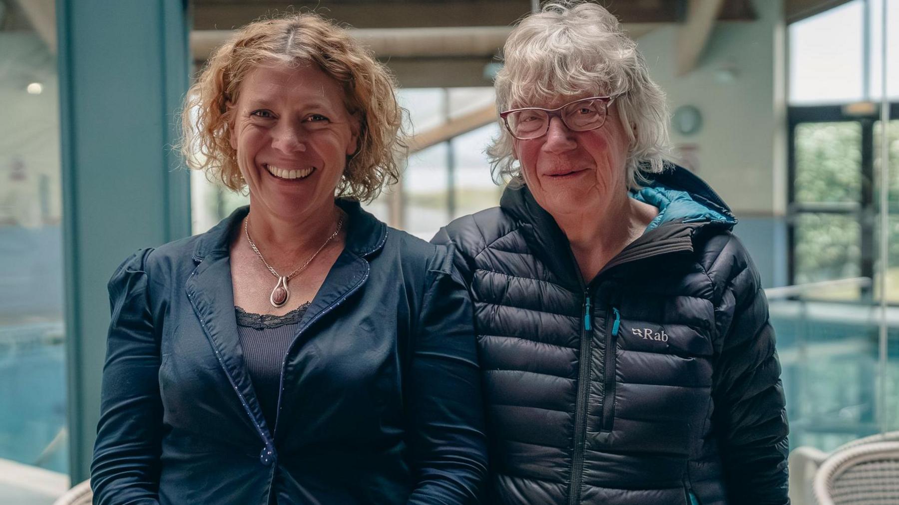 Two women stand facing the camera smiling. The women on the left has short curly ginger hair and is wearing a navy jacket. The woman on the right has short grey hair, glasses and a dark coat. 