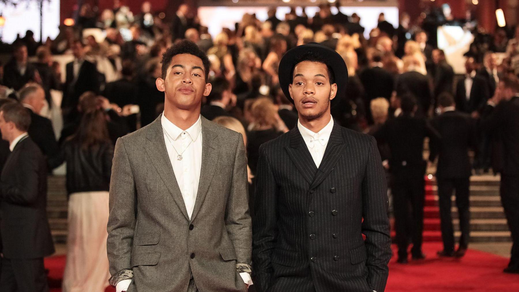 Jordan and Harley from Rizzle Kicks are pictured wearing suits at a film premiere. They both have buttoned-up white shirts with no ties. Jordan is wearing a grey suit while Harley wears a black pinstripe suit with a hat. They are both looking serious and their eyes are focused slightly off-camera.