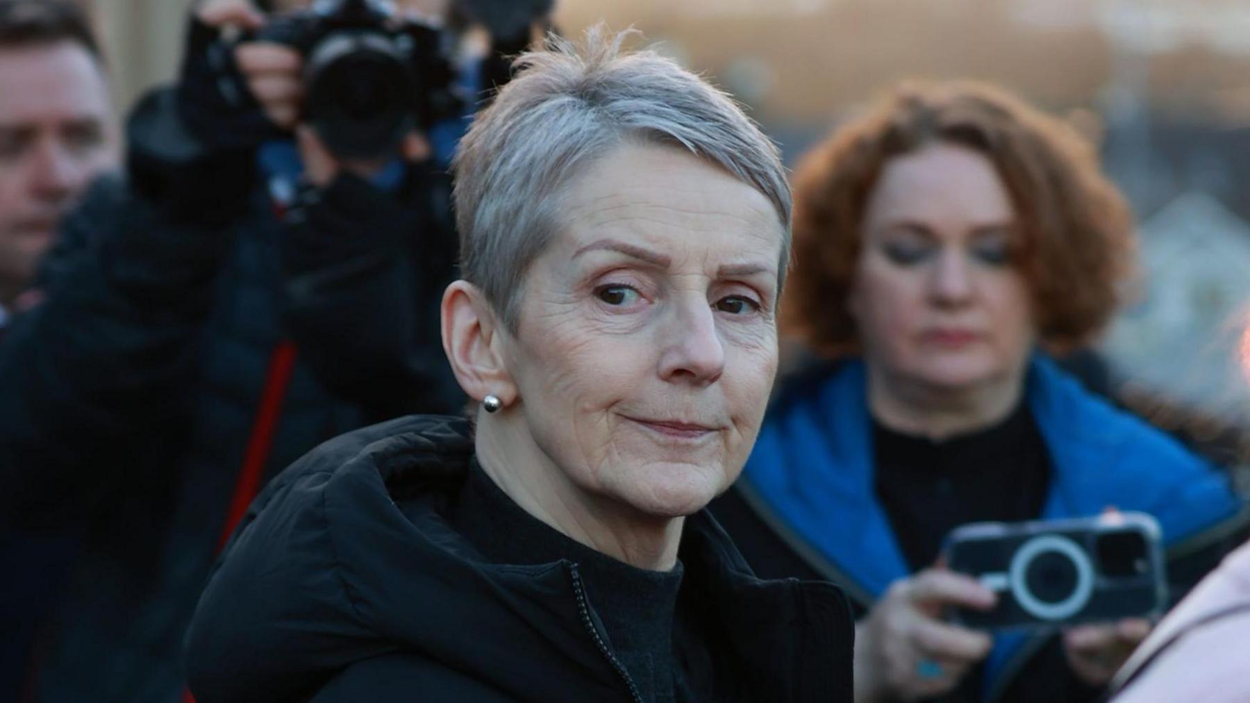 A close-up image of Lady Eleanor Donaldson outside court in Newry. Behind her are reporters with cameras and phones pointed at her. Lady Donaldson is an older woman with short grey hair. She has a neutral expression on her face. She is wearing a large, black coat.