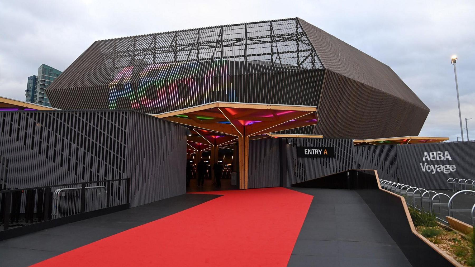 An exterior shot of the ABBA Arena in London showing a red carpet leading up to the entrance. The word "ABBA" can be seen in the pattern of the metalwork of the roof