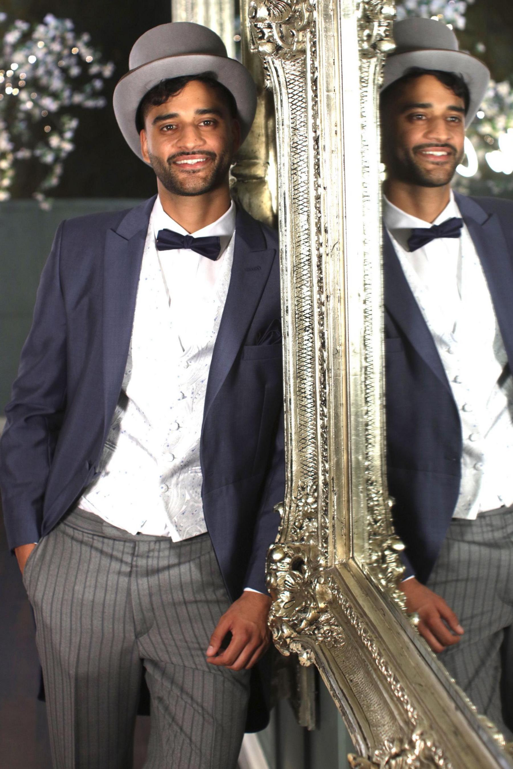 Bal leans against a wall in front of an ornate silver-framed mirror, in which he is reflected in his top hat and tails.