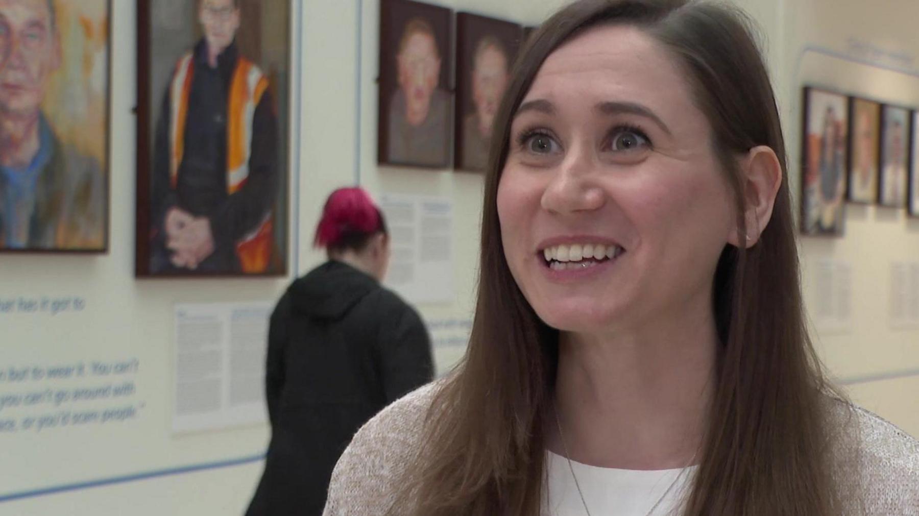 A youngish woman with straight brown hair is smiling to the left of the frame. She is wearing a white top, beige cardigan and a chain necklace. Behind her is a wall of portraits. A woman in a black jacket and with pink hair in a ponytail is walking away in the background.