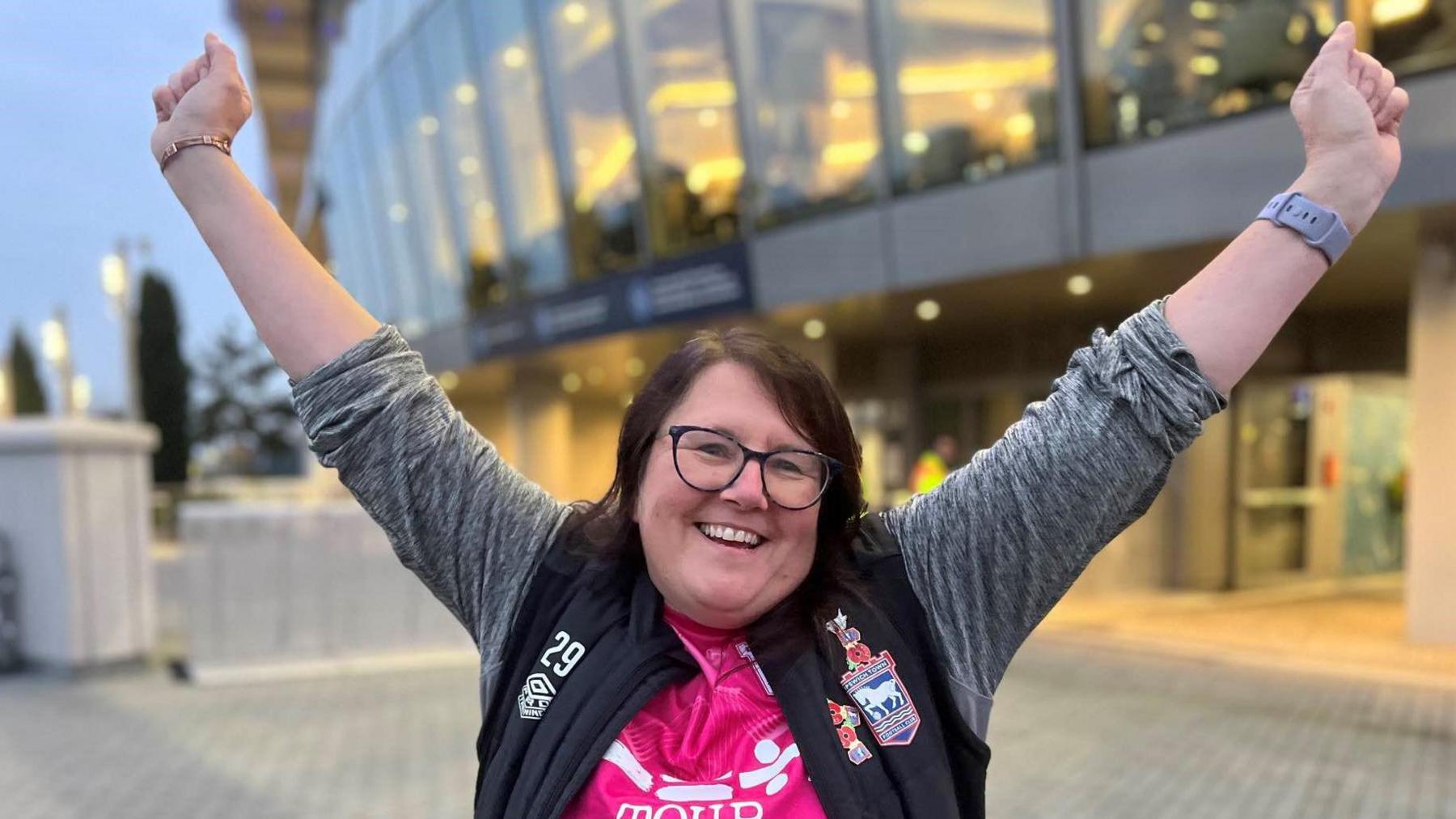 A woman wearing glasses with shoulder length dark brown hair with a side parting.  She is wearing the pink Ipswich Town away shirt covered by an open long sleeved jacket with a black body and grey arms.  The jacket has a large Ipswich Town logo on it.  She has her arms aloft in a show of victory.  In the background is the exterior of the Tottenham Hotspur stadium.  