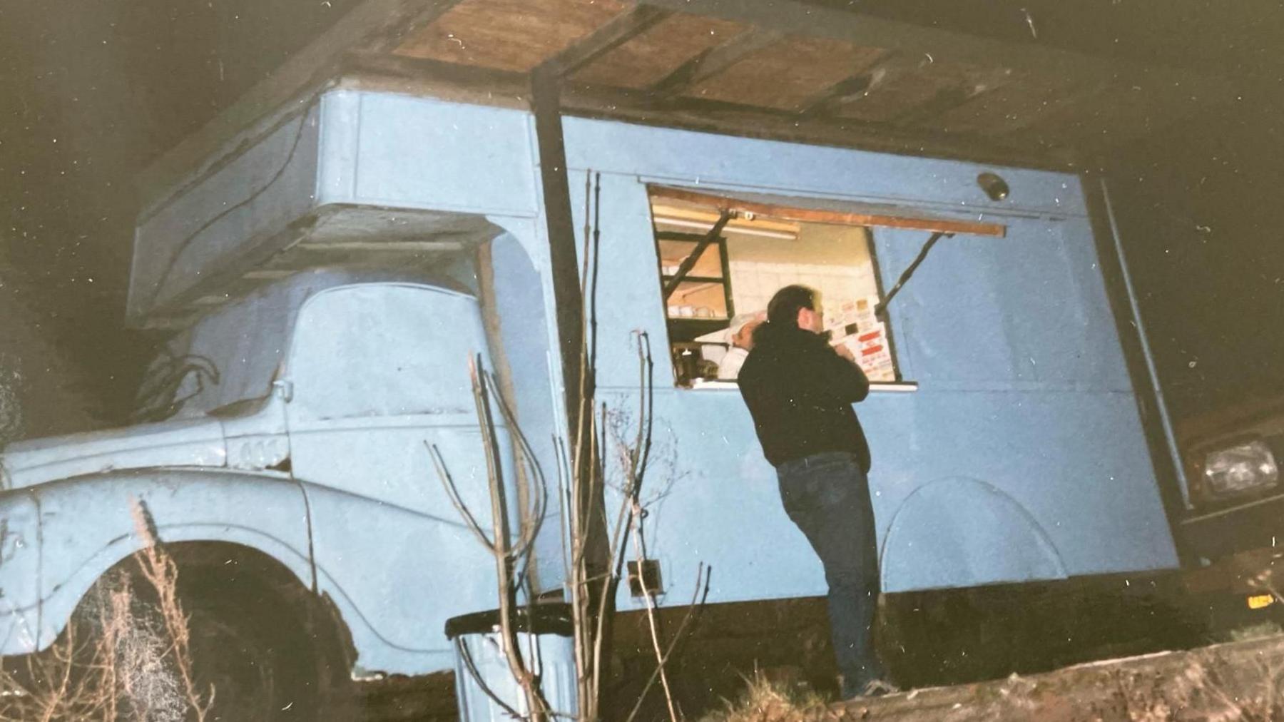 A man in a black jacket stands in front of a pale blue food truck