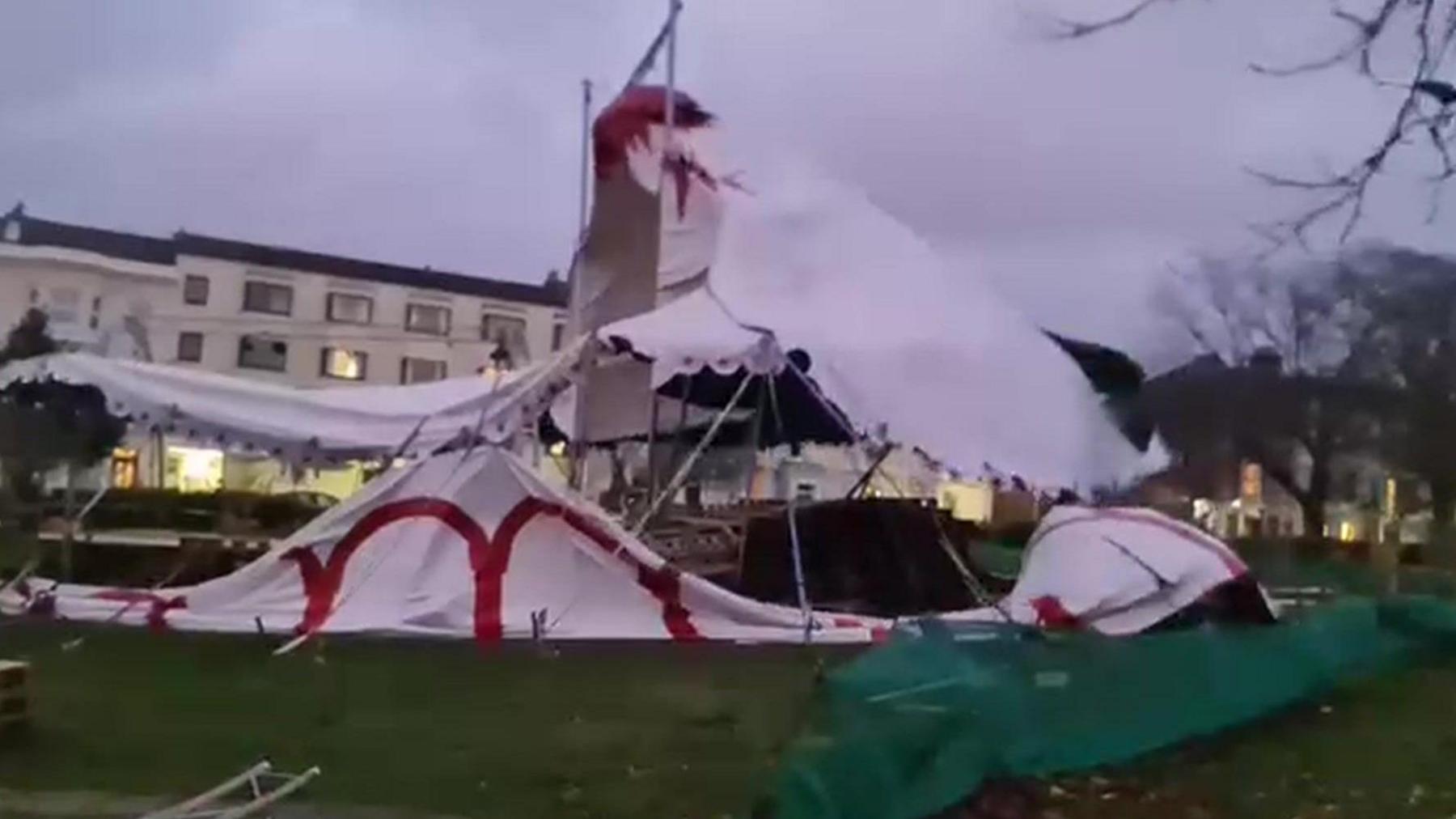 A large white tent with a big rip and red markings in a park with buildings behind.