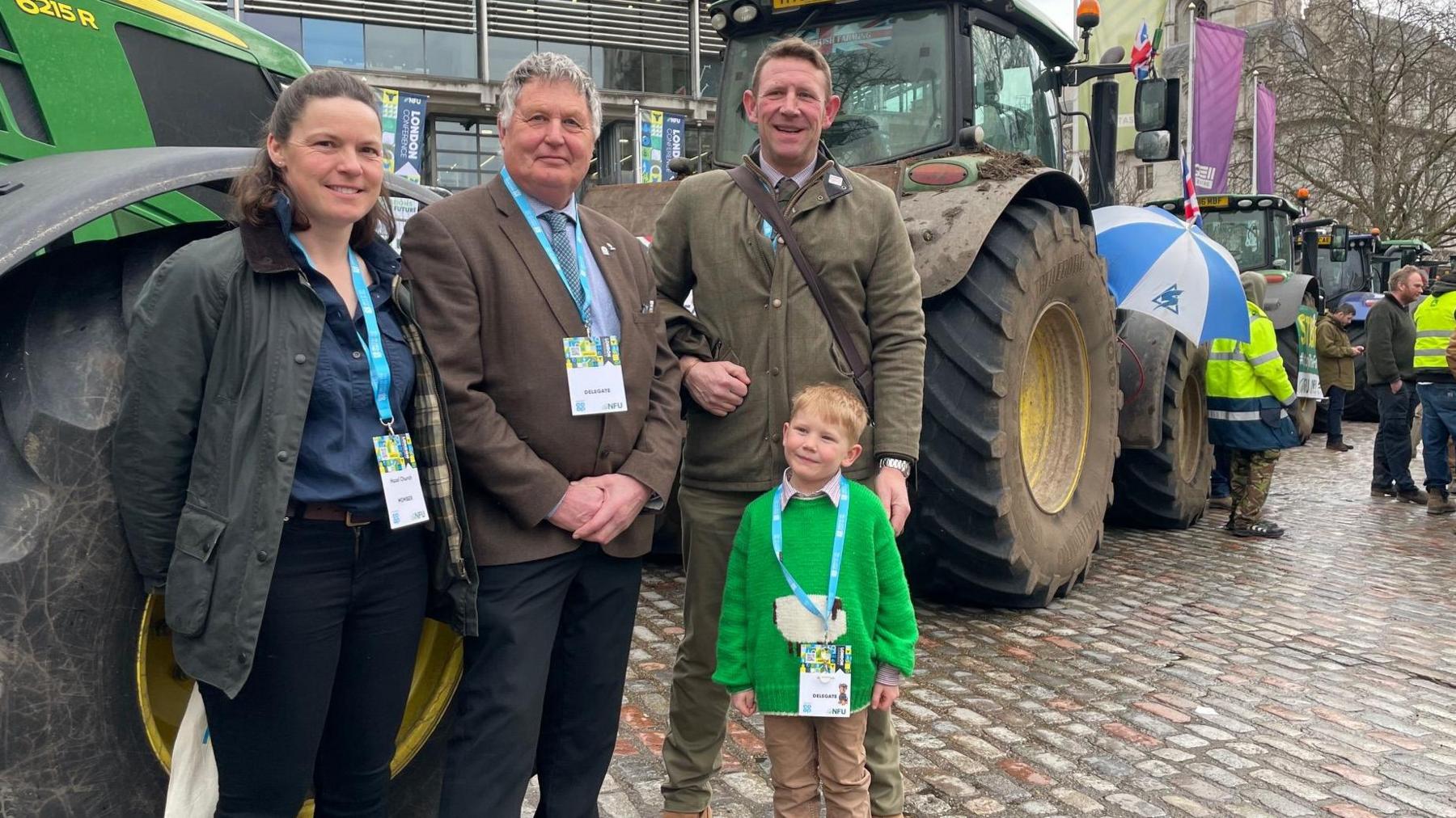 Hazel and Tom Church with Martin Towler, and five-year-old Bertie, standing by a line of tractors in a wet cobbled street. Other farmers stand in the background in hi-viz jackets