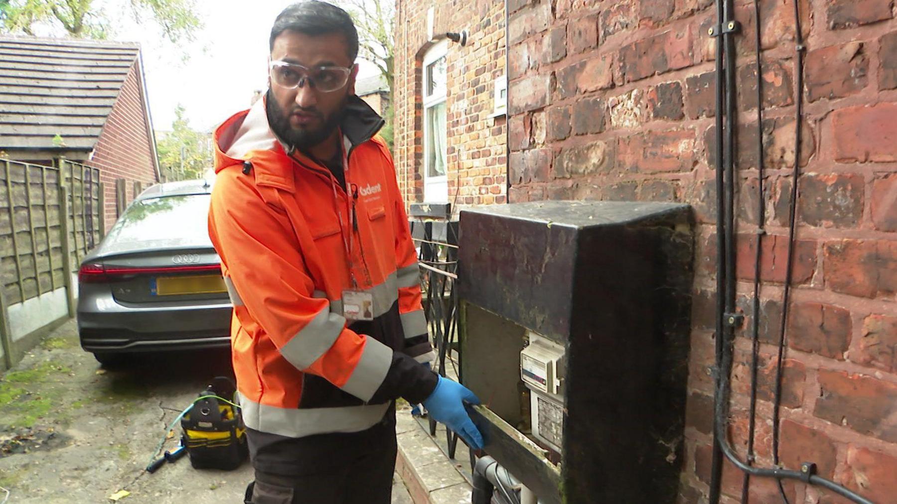 Asif Hussain is stood next to a gas meter at the side of a house. He is wearing an orange Cadent jacket, blue rubber gloves and safety goggles.