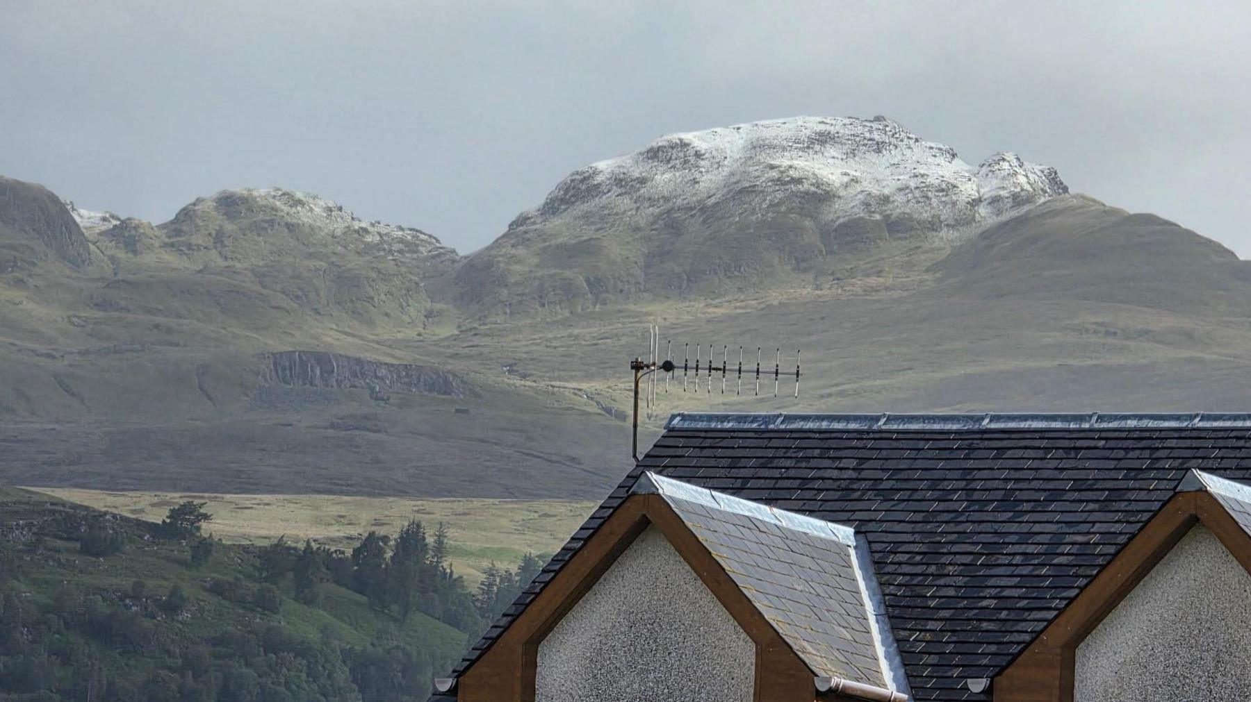 Looking out over the roof of a house are lumpy green hills with a dusting of snow.
