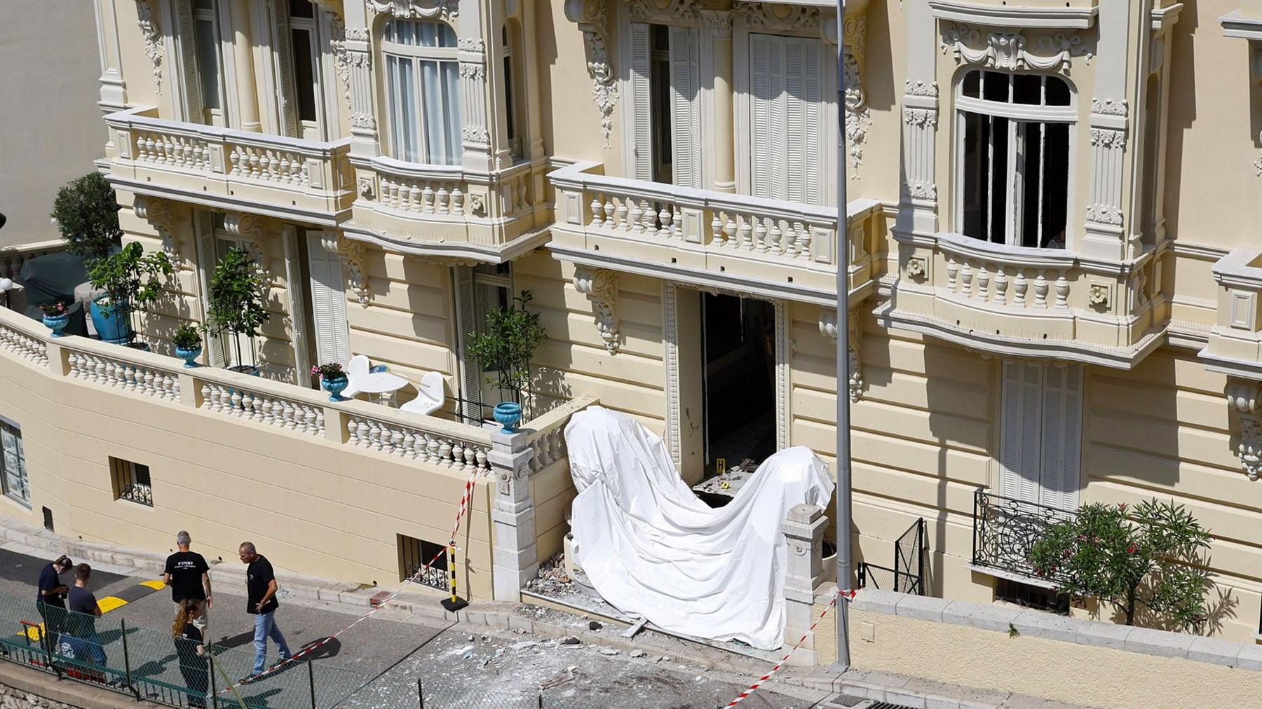 Frontage of a large cream coloured residential building in Monaco with damage to windows and a white sheet accross the front steps and police standing near the front of the building