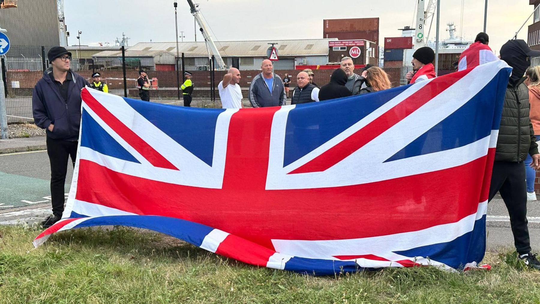 Two men, one of them masked, holding up a large union flag with a ferry port in the background