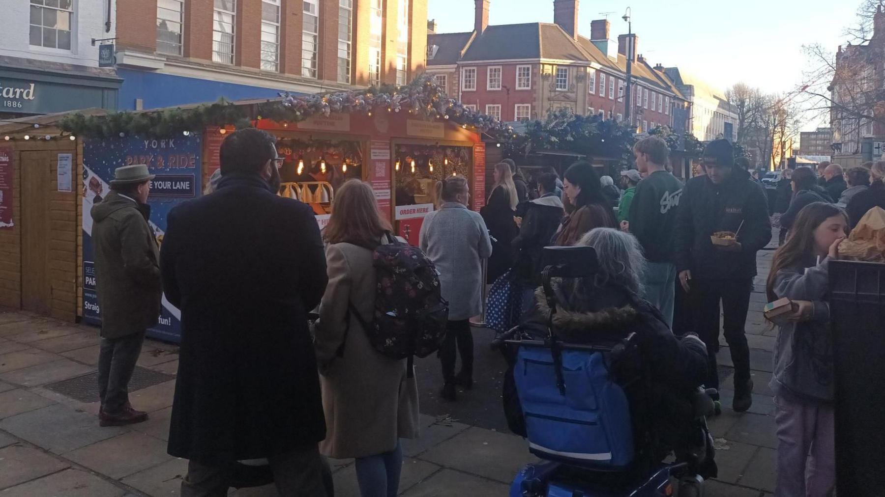 A shot of the Christmas Market in York, with a large fir tree archway, decorated with gold, green and red baubles and illuminated stars. A mixed crowd of people can be seen walking beneath it.
