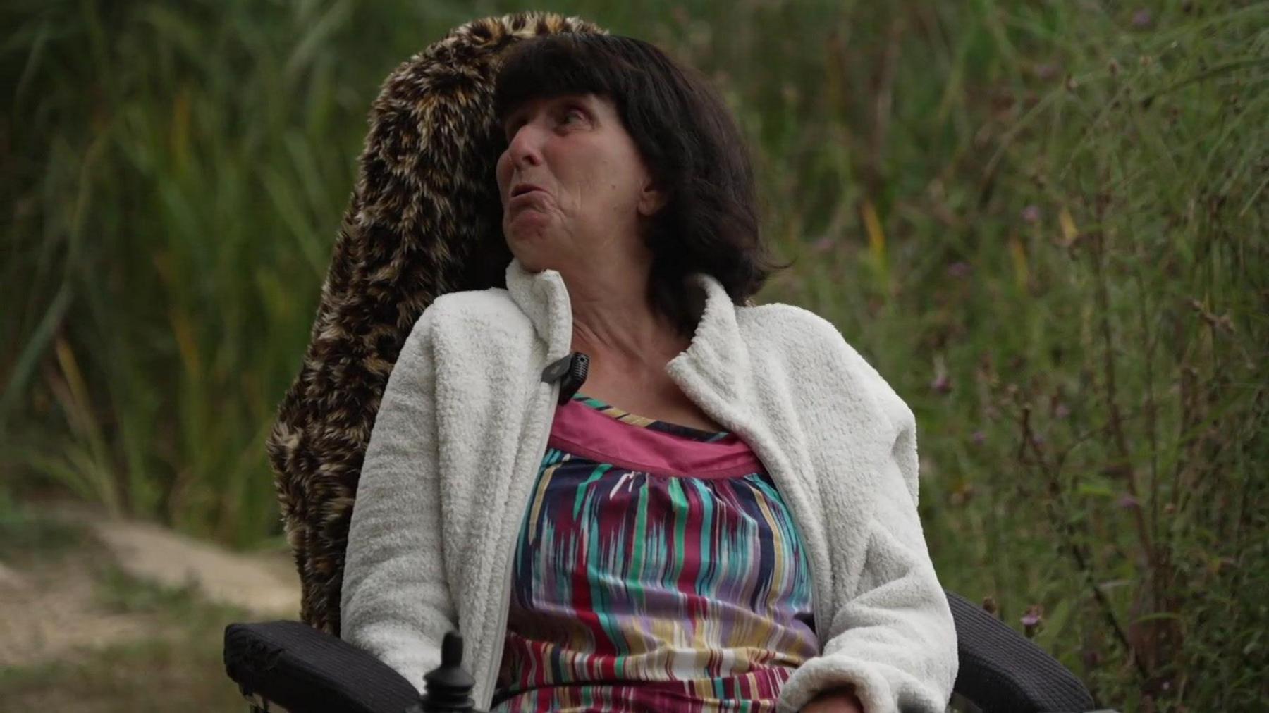 Carol Pop sits in her wheelchair on the bank at Southampton Common. In the background behind her are tall green reeds which line the lake. Her chair has a tall back, which has been covered in a fur leopard print cover. Carol has a short dark brown bob hairstyle with a fringe. She wears a multi coloured abstract stripey blouse with a white unzipped fleece over the top. She is looking to the side as if she is talking to someone off camera.