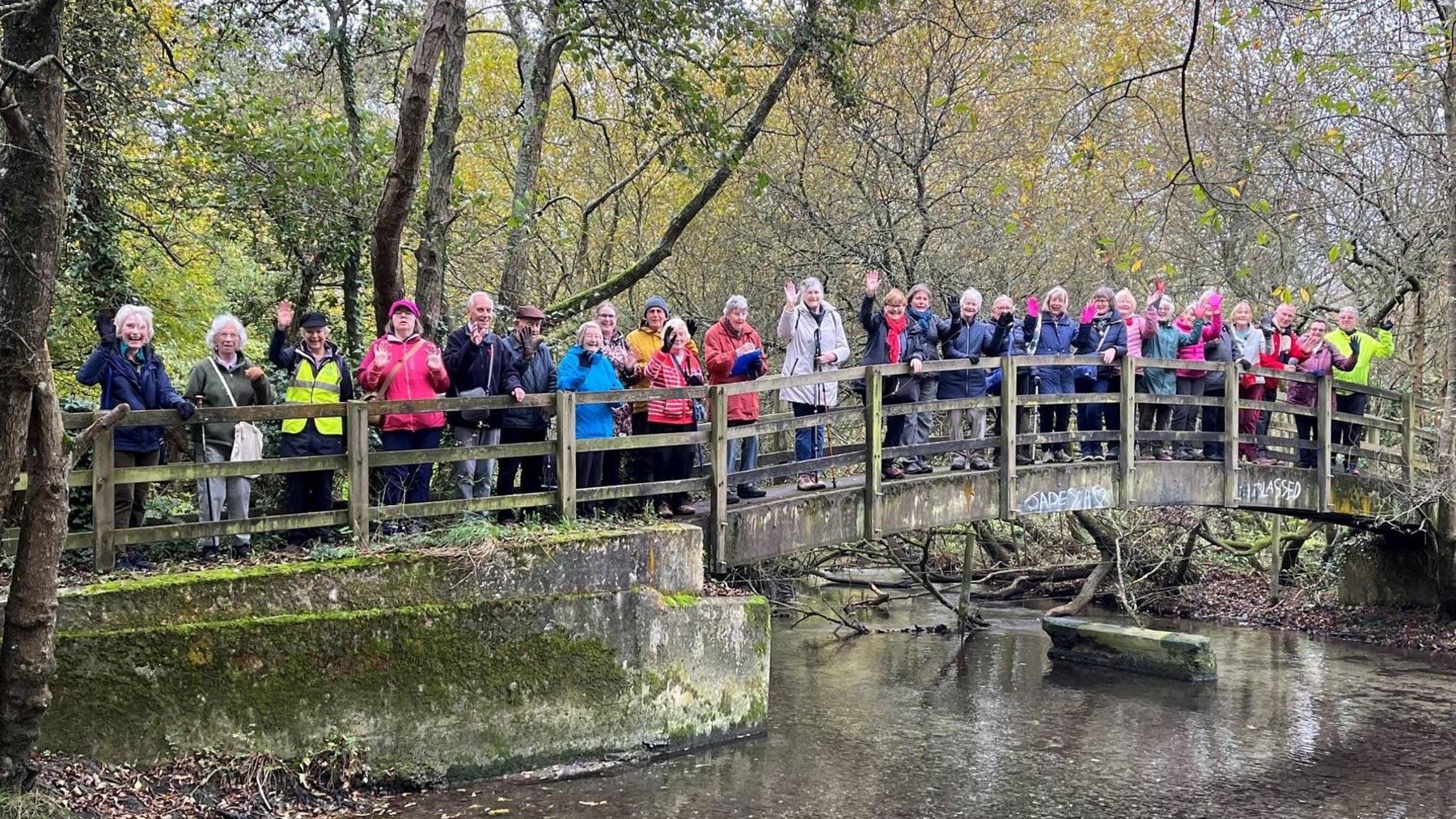 A large group of walkers, wearing different coloured coats, standing on wooden footbridge over the River Nadder.