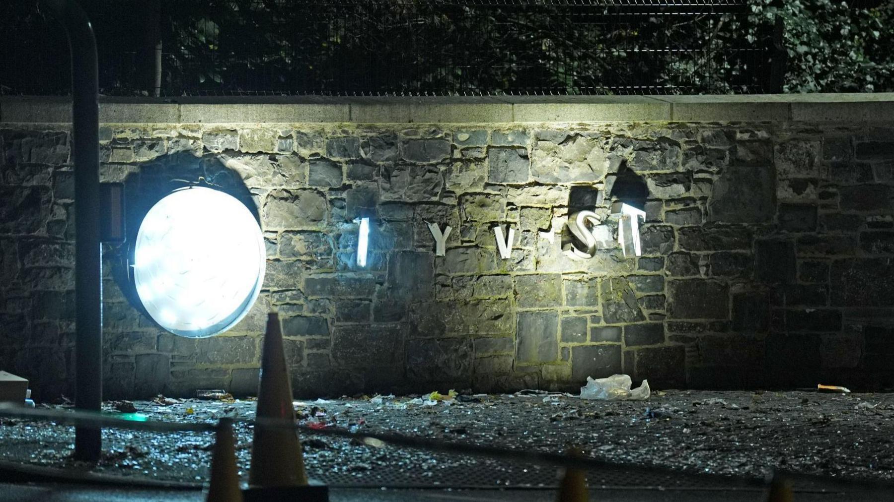 Silver letters are on a stone wall, some have fallen of. The wall is a collection of coloured stones with a black fence on top. In front of the wall there is a collection of rubbish. There are orange cones in front of the rubbish. 