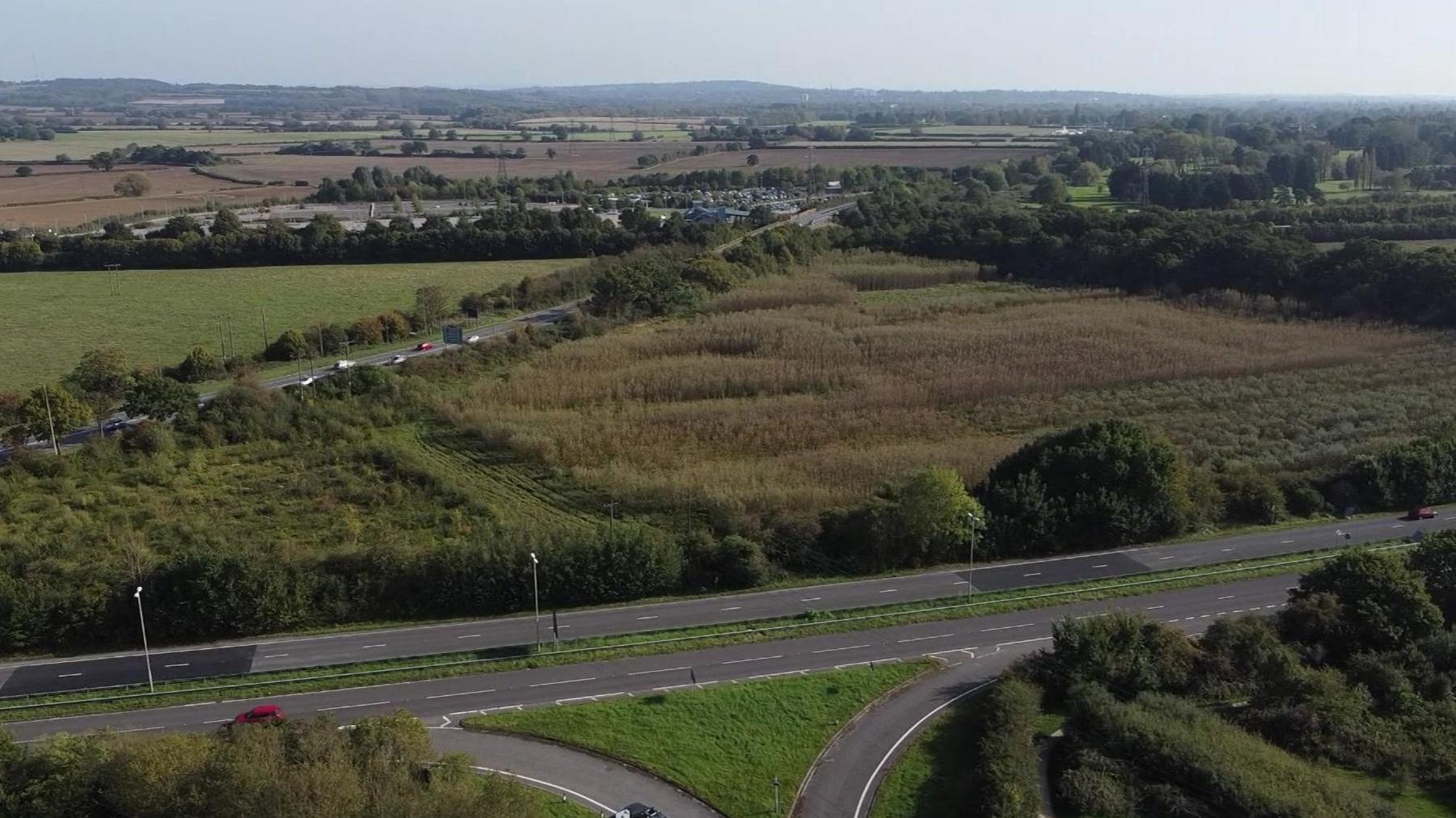 Drone image of a triangular-shaped piece of green land with roads to the bottom and to the left and fields beyond.