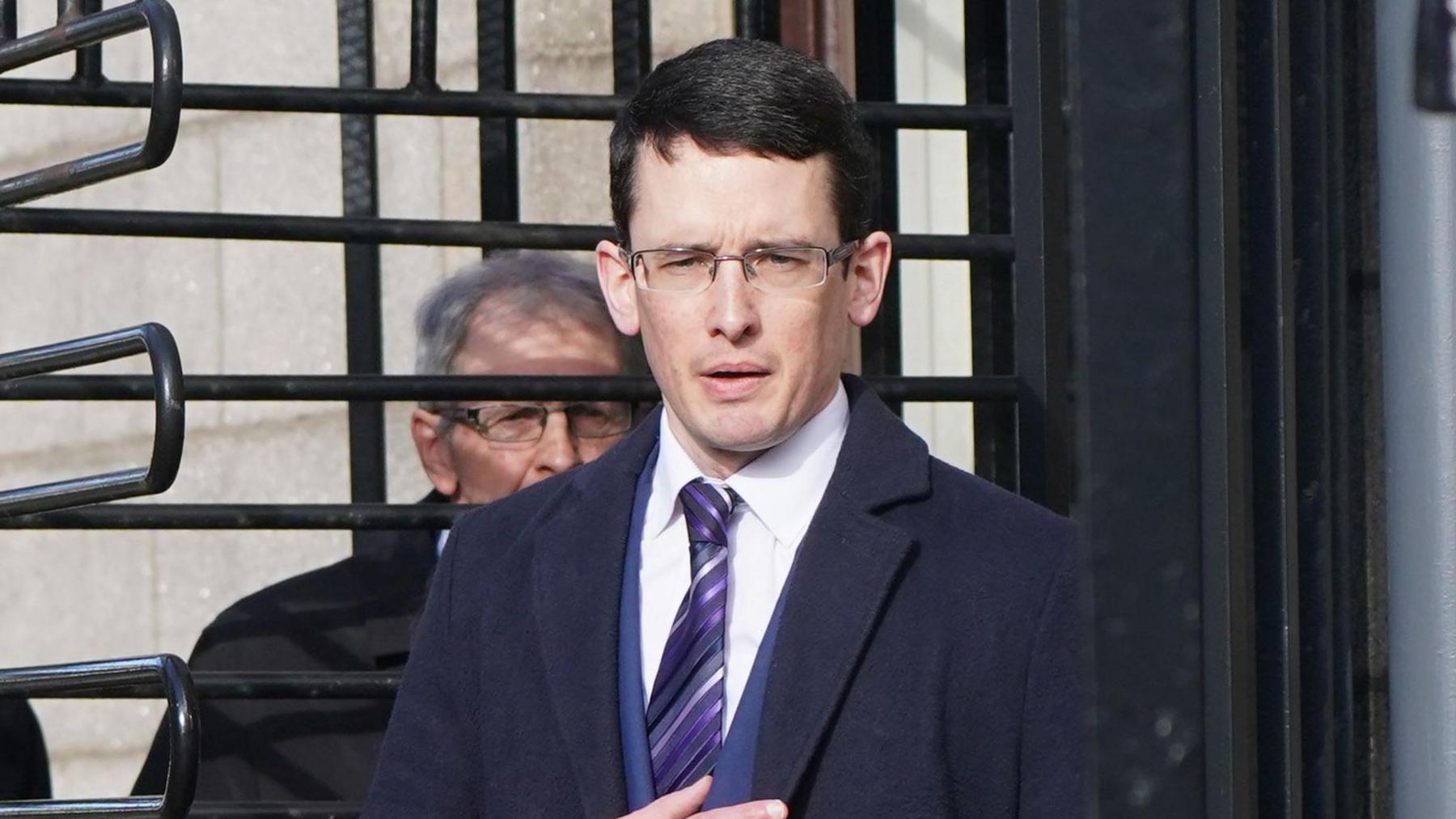 Enoch Burke, wearing a suit and tie, walks through a turnstile