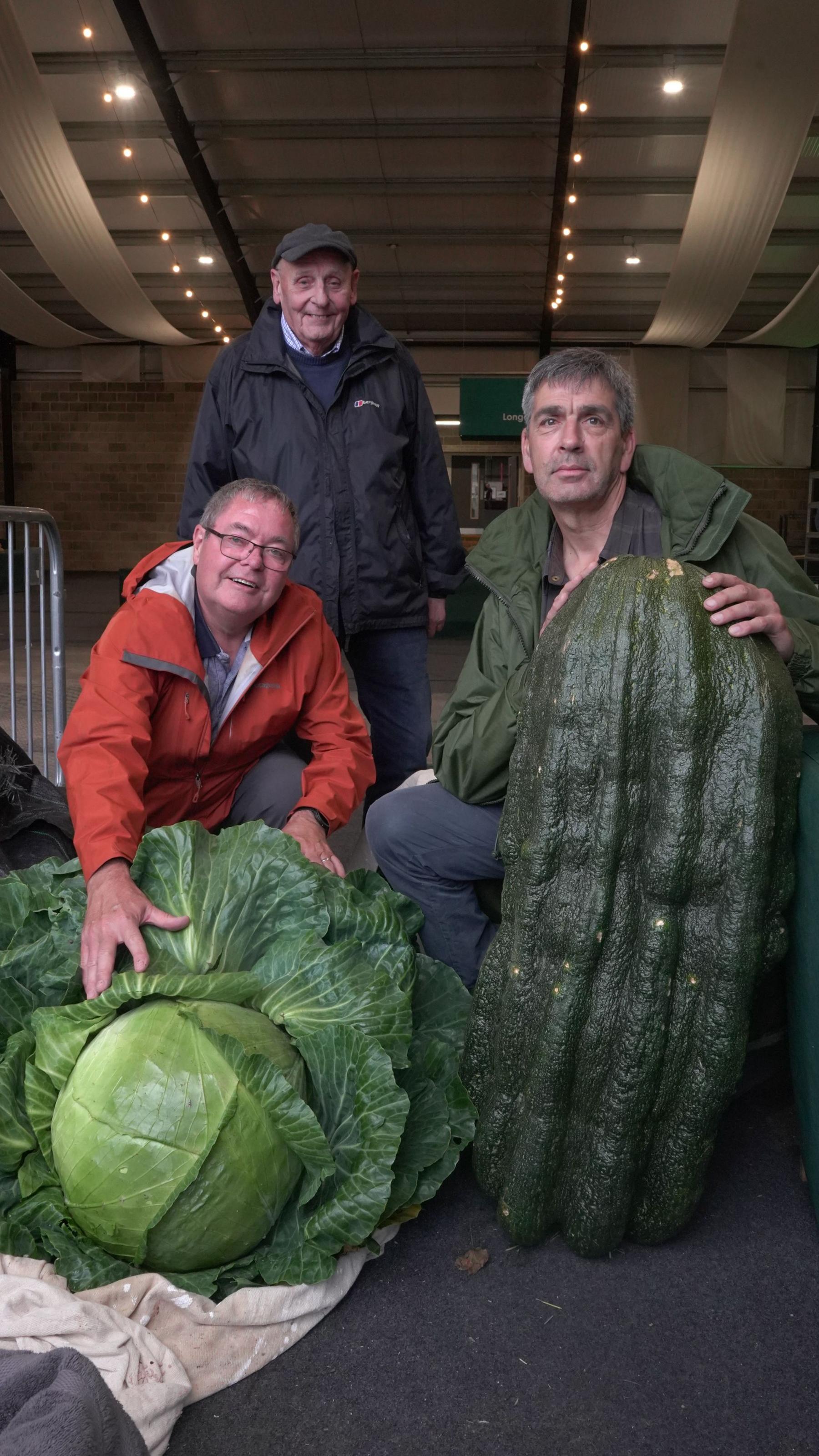 Three men pose with a huge cabbage and a huge squash. 