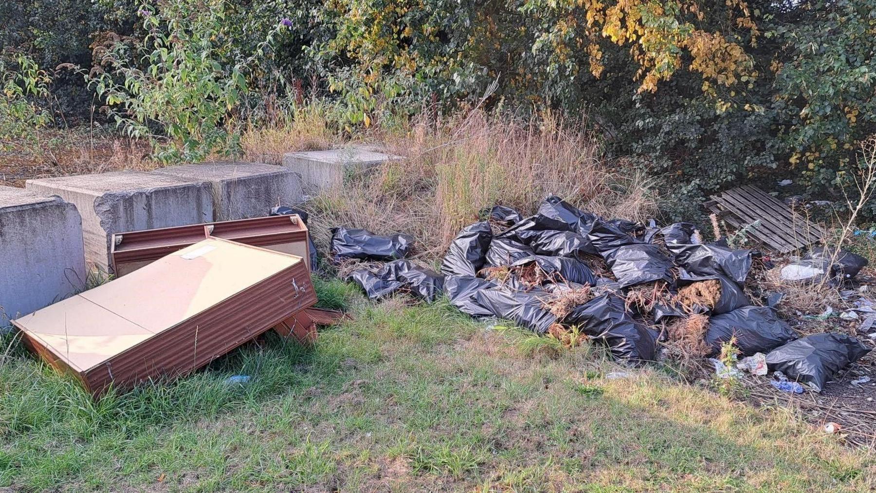 There is a large pile of black bin bags next to broken brown furniture that has been dumped on a grassy area. 