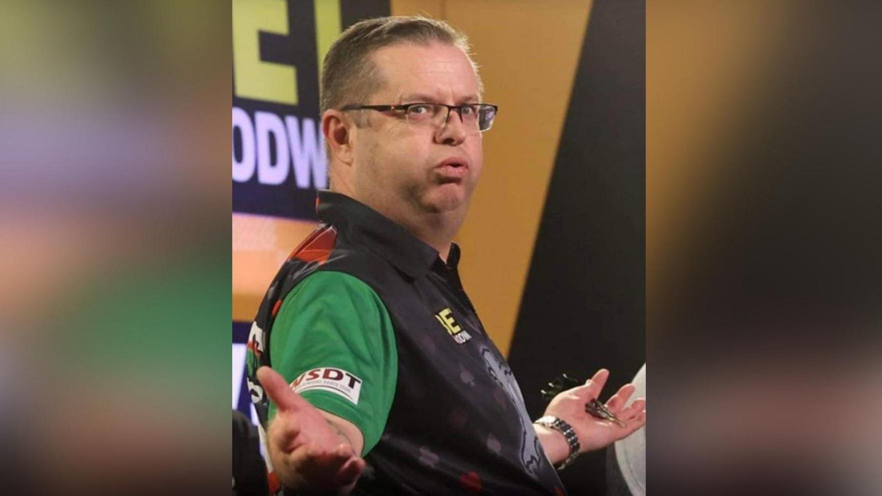 Llun Richard-Eirig Rowlands yn sefyll gyda'i ddwylo allan. Mae'n dal 3 dart yn ei law chwith. Mae ganddo wallt brown byr a sbectol. Mae'n gwisgo crys-t du a gwyrdd.