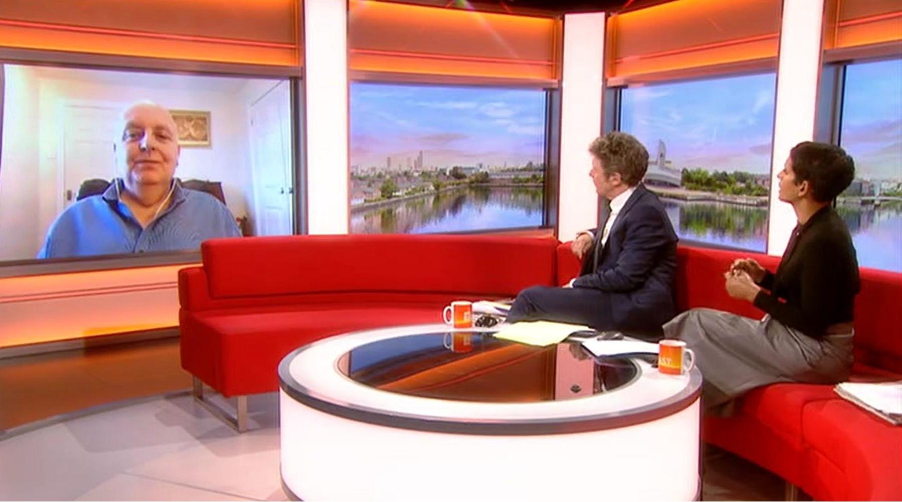 A man (Charlie Stayt) and a woman (Naga Munchetty) sitting on a red sofa in a studio, with a white circular table in front of them. They are looking at Mark Morrell, who is on a video screen wearing a blue shirt