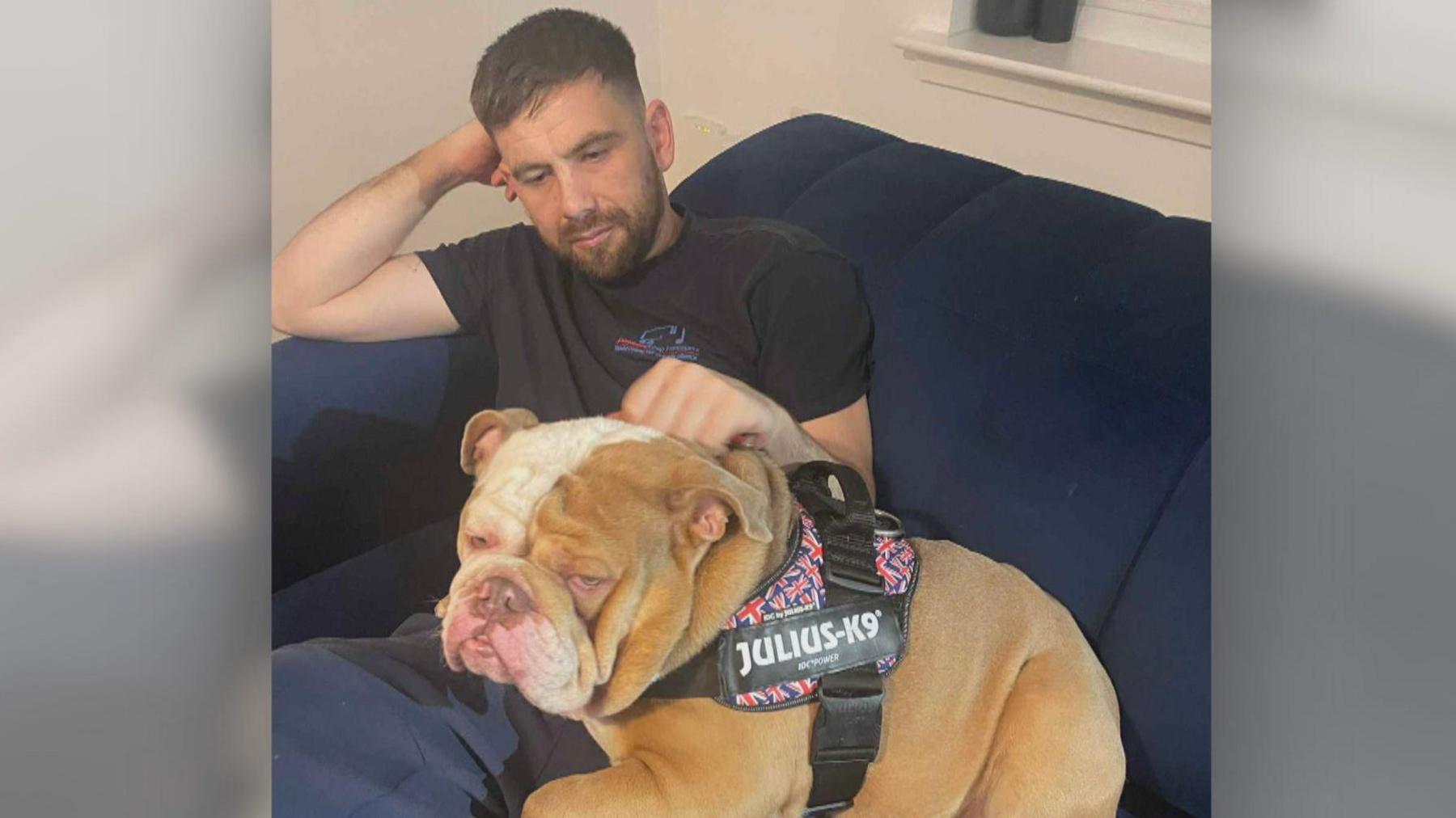 Greg Monks wearing a black t-shirt with his right arm raised. He is sitting on a sofa with a large brown and white dog wearing a black harness sitting on him.