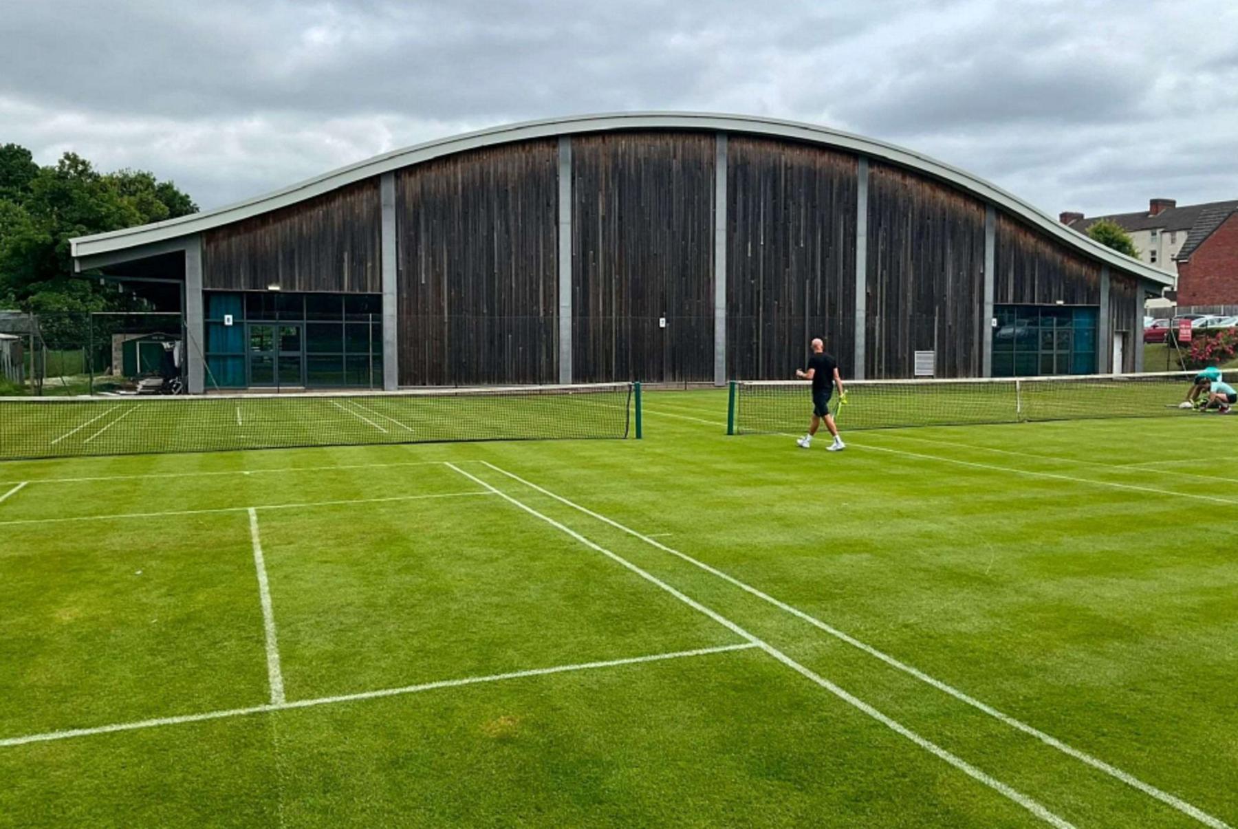 Two outdoor grass tennis courts. A man wearing black shorts and a black t-shirt is walking towards a gap between the two courts, while holding a racket. A large wooden building with a curved silver roof is in the background.