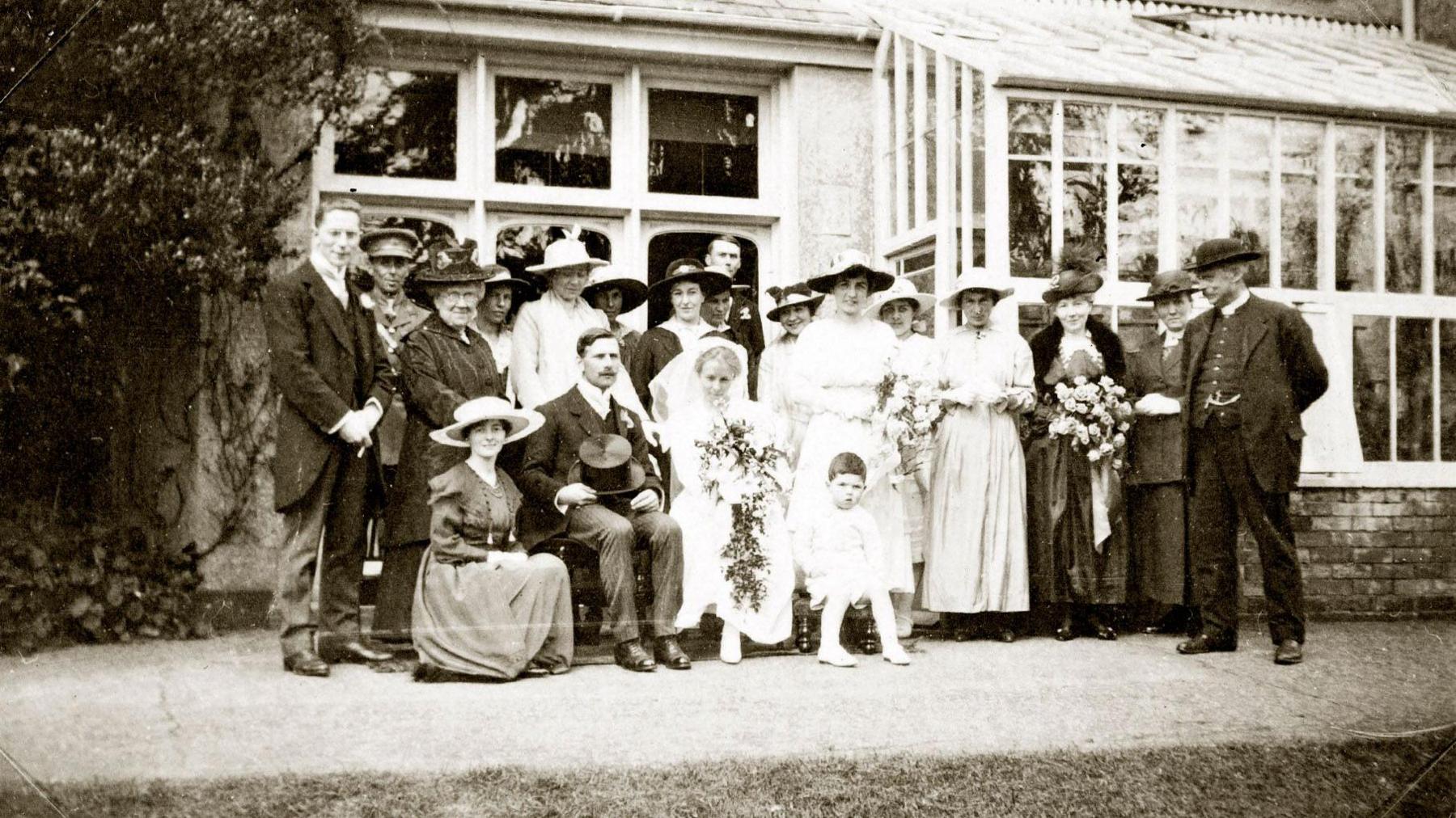 A black and white image showing a group of around 20 adults, standing up, in old-fashioned formal clothes, including wide-brimmed hats, long dresses and suits. There is a couple sitting in front of them, with a man holding a top hat in his lap and a woman in a wedding dress. She and several of the standing women are holding large floral bouquets. Behind the group is an orangery with many glass panes. 