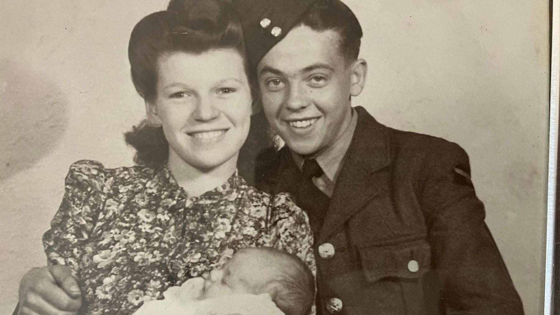 Tommy Doherty is on the right of the image taken in the 1940s, with his wife to the left. They are both smiling in the black and white photo. She is holding a bay, and is wearing a patterned dress and with dark hair. Tommy is in military uniform, and has his arm around her. The baby appears to be asleep and is wrapped in a light coloured blanket.