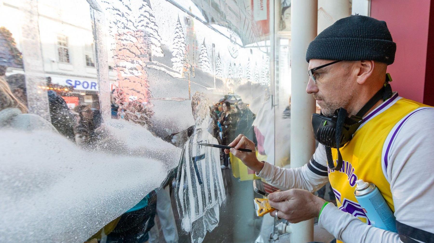 Man with yellow and white top wearing a beanie hat to the right of a window sprayed with decorative show - he is working on a snow scene with trees.