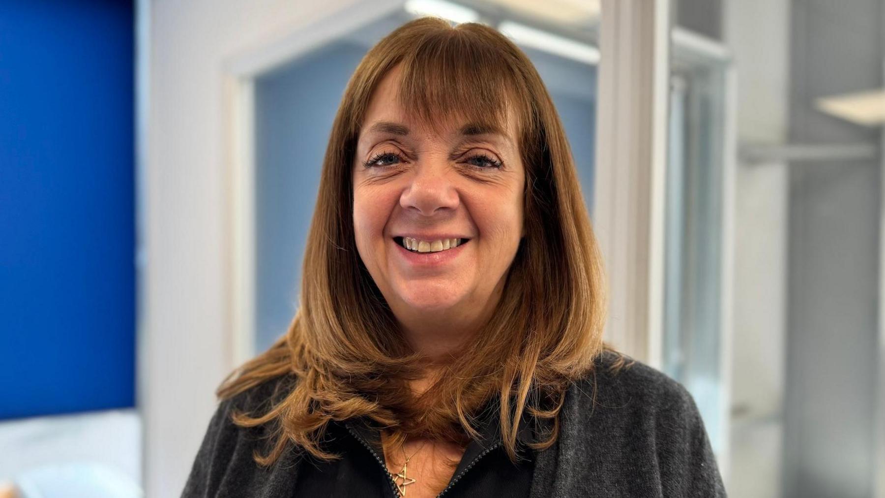 Jackie Betteridge has medium length brown hair that curls at the ends, and has a fringe. She is smiling, wearing a grey top and standing in front of office walls with windows and a blue blind to the left.