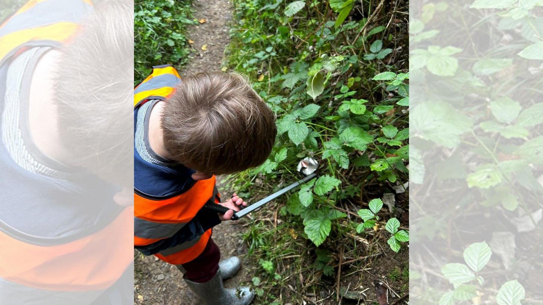 a young boy in a high vis jacket holding a litter picker reaches for a crushed can caught in a plant just off a woodland trail