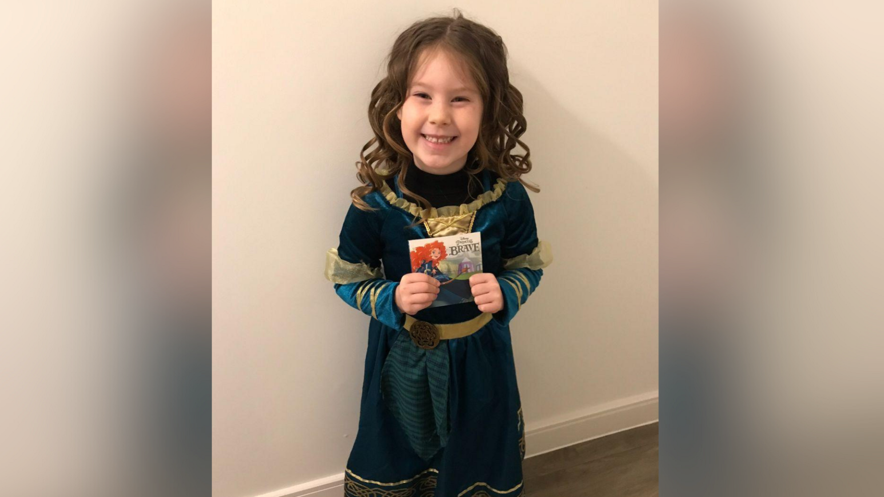 A child dressed up for World Book Day wearing a green dress and holding a book. She is stood in front of a white wall. 