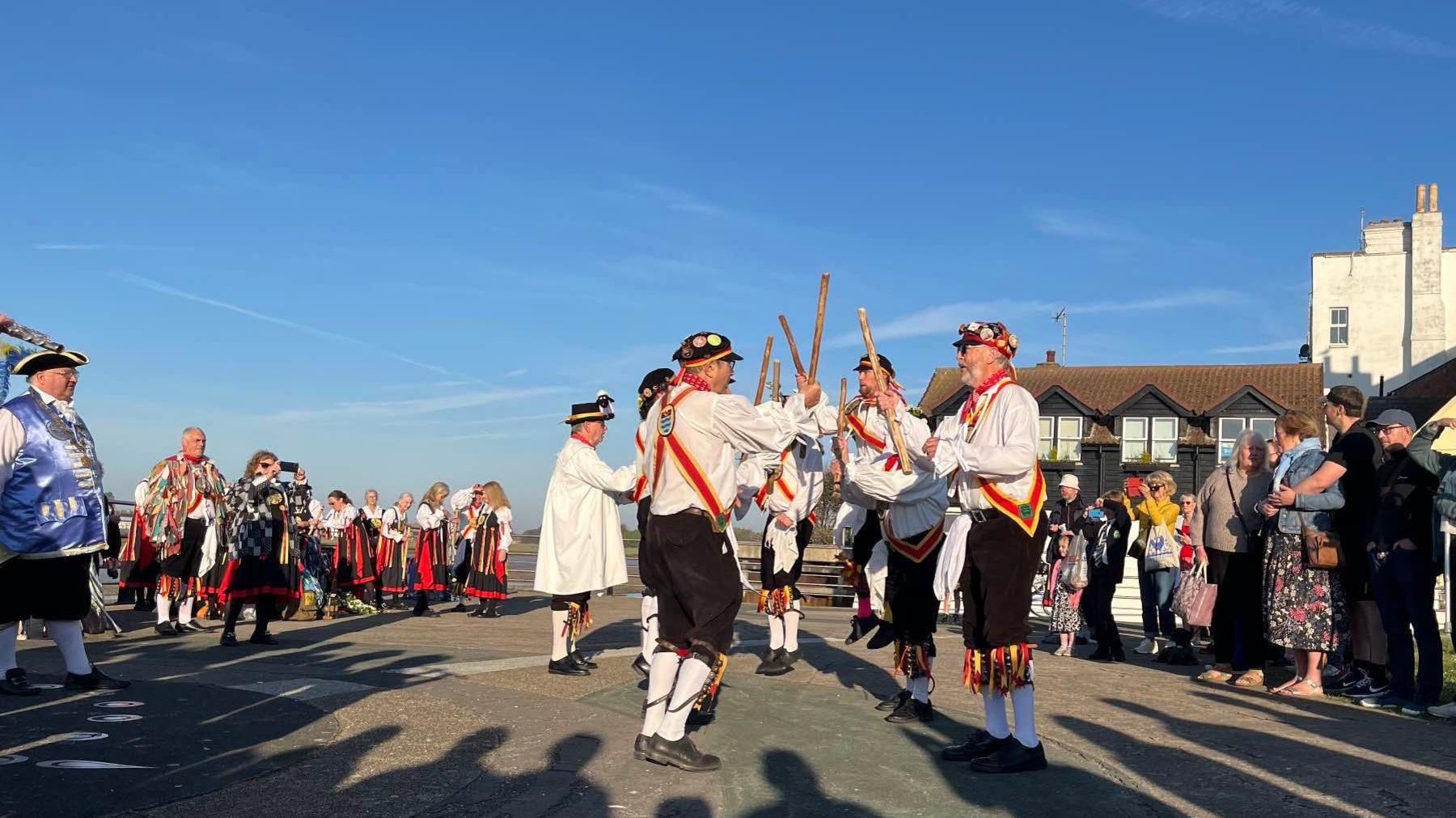 Morris dancers in the traditional dress of white shirts and black trousers with red and yellow ribbons dance in formation opposite each other and hold their sticks in the air. Behind them is another Morris group and the sky above is blue.