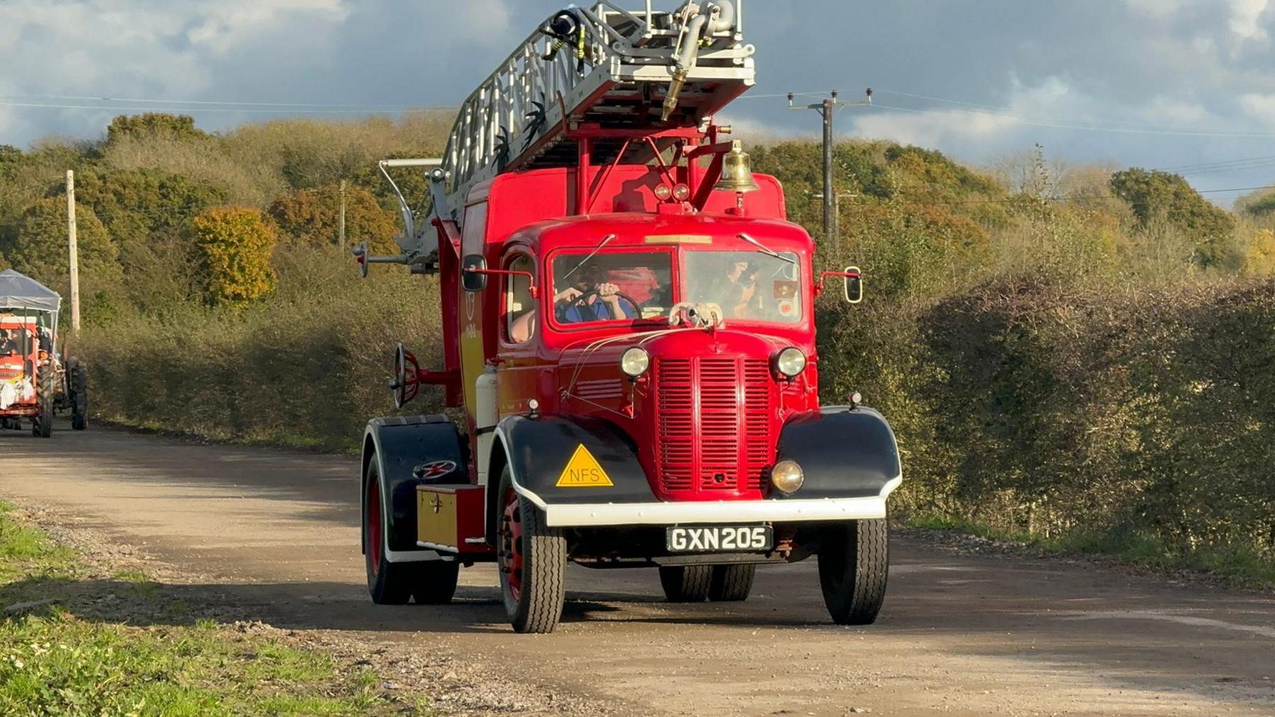 A classic red fire engine with a bell and a ladder on its roof drives down a country lane. 