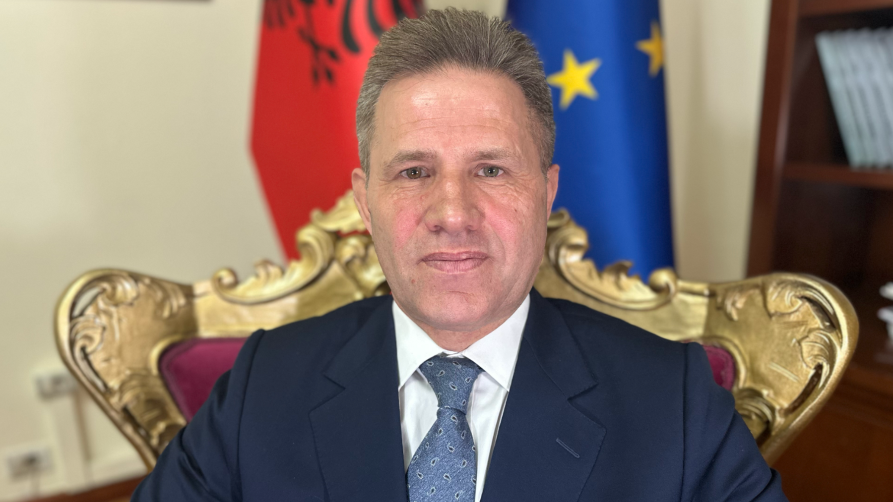 Lavdrim Krashi sitting inside the government building in Tirana, the Albanian capital, on a chair in front of flags for Albania and the European Union