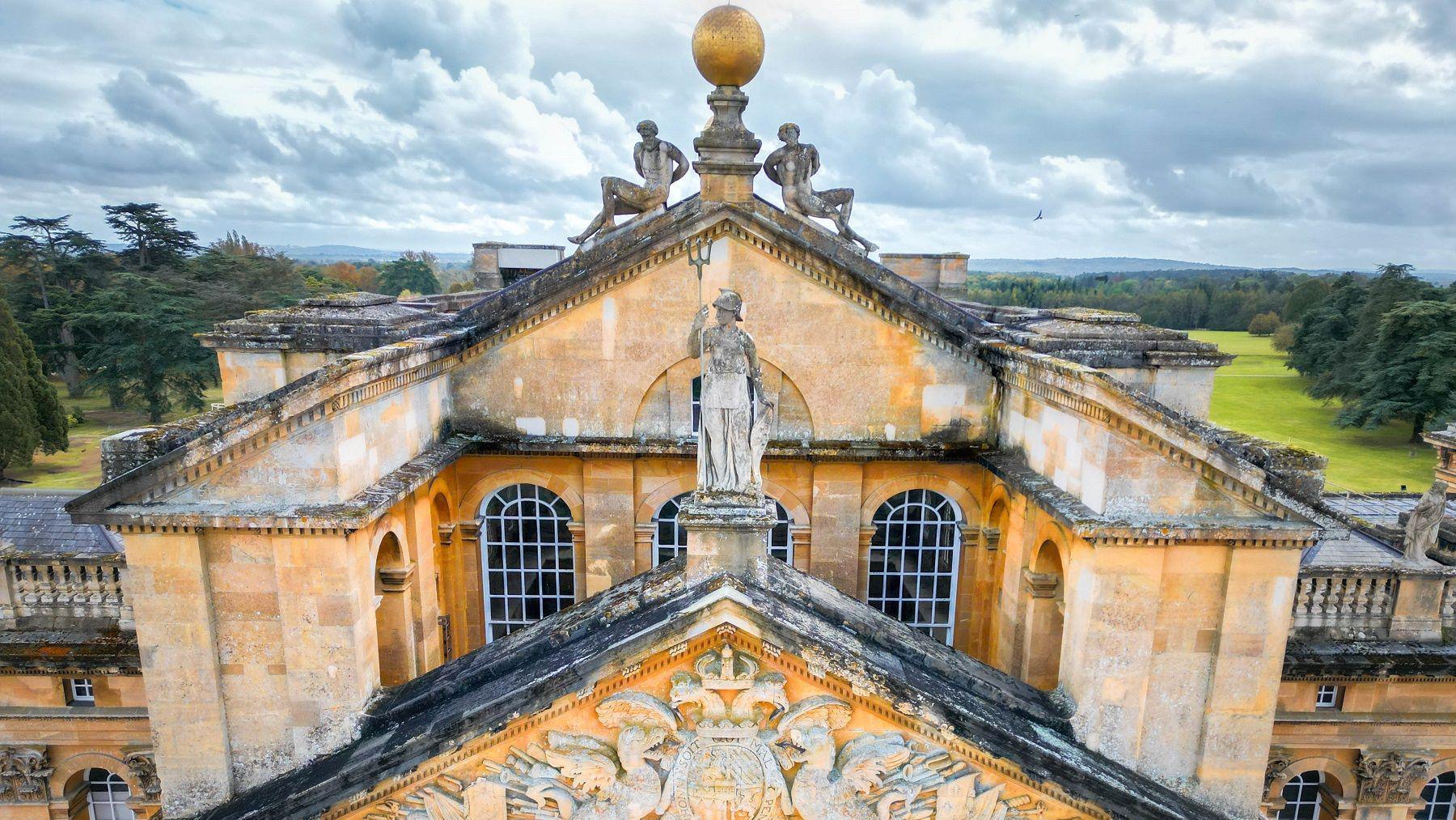 An aerial view of part of the roof at Blenheim Palace. There are statues and ornaments on top of it. The palace's grounds can be seen in the background. It's cloudy.