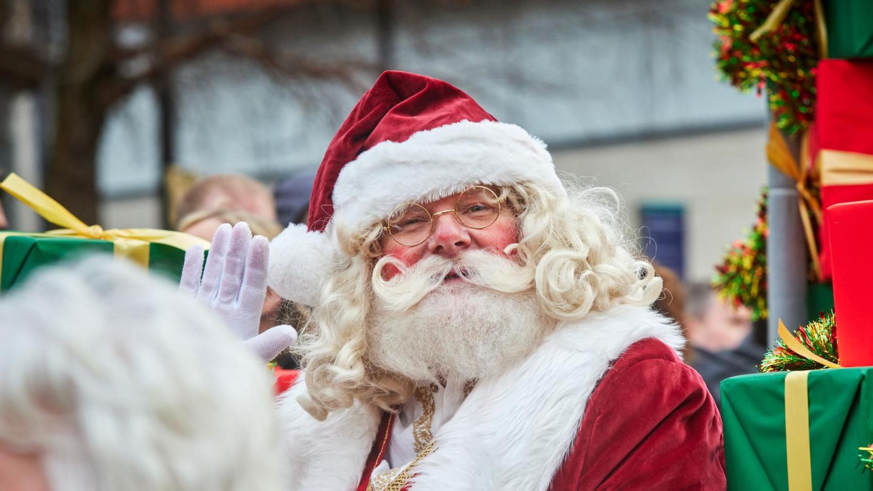 Santa Claus waves to the camera surrounded by wrapped gifts