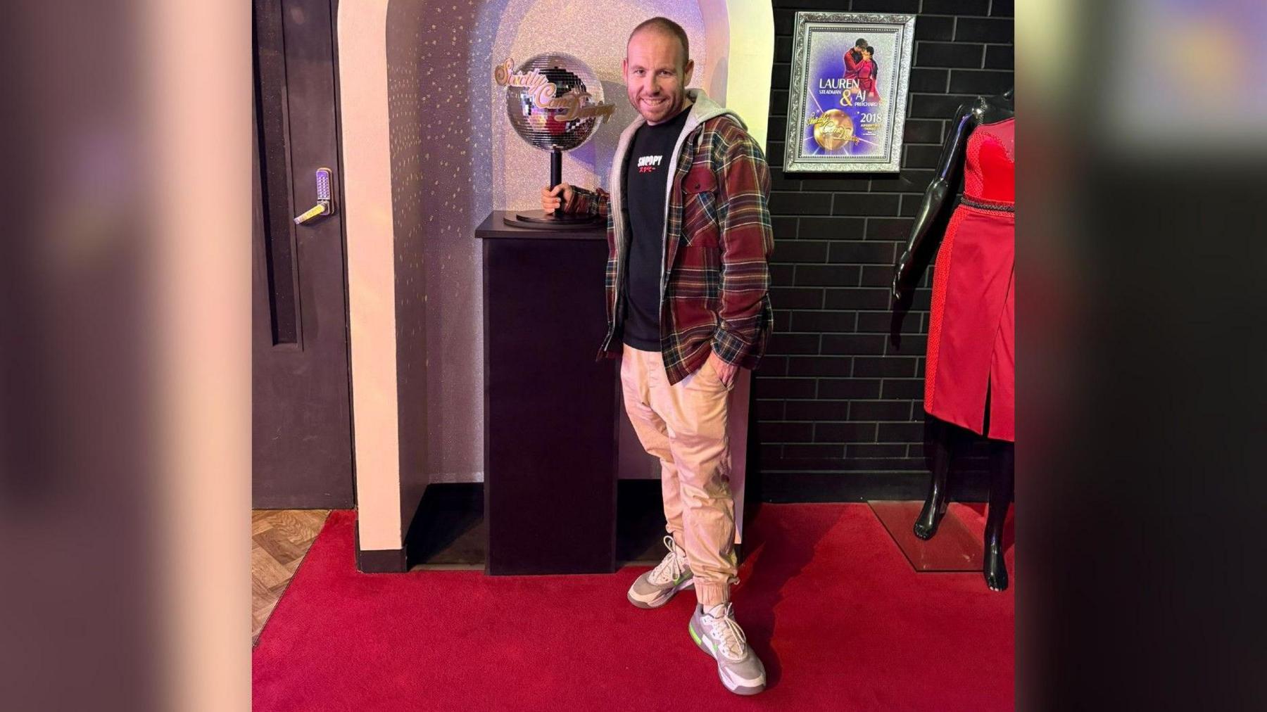 Lee Gigg stod on a read carpet next to the Strictly Come Dancing glitterball wearing beige trousers and a plaid jacket