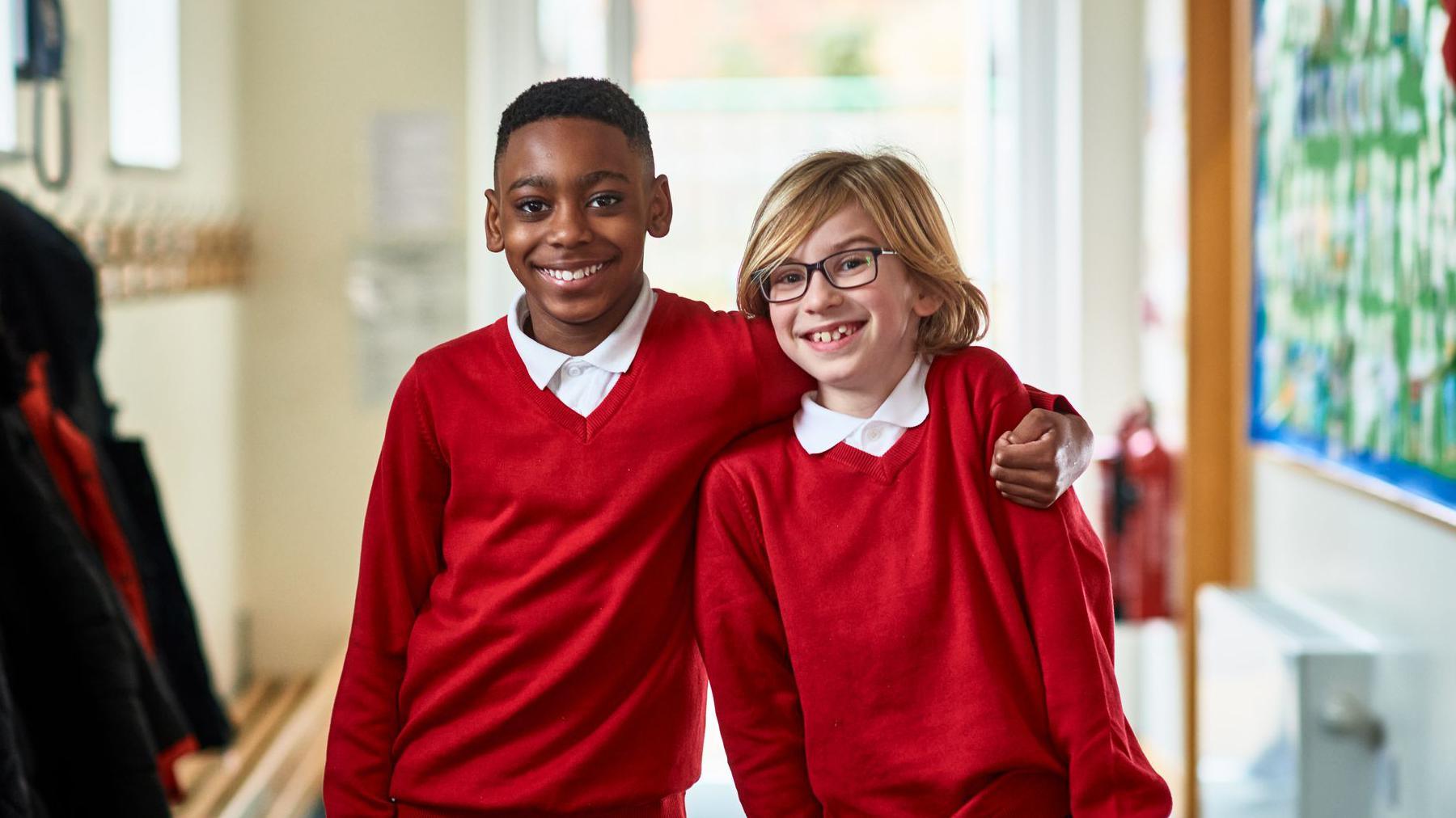 Two boys stand in a school corridor. One has their arm around the other's shoulders. They are wearing white polo shirts and red jumpers.
